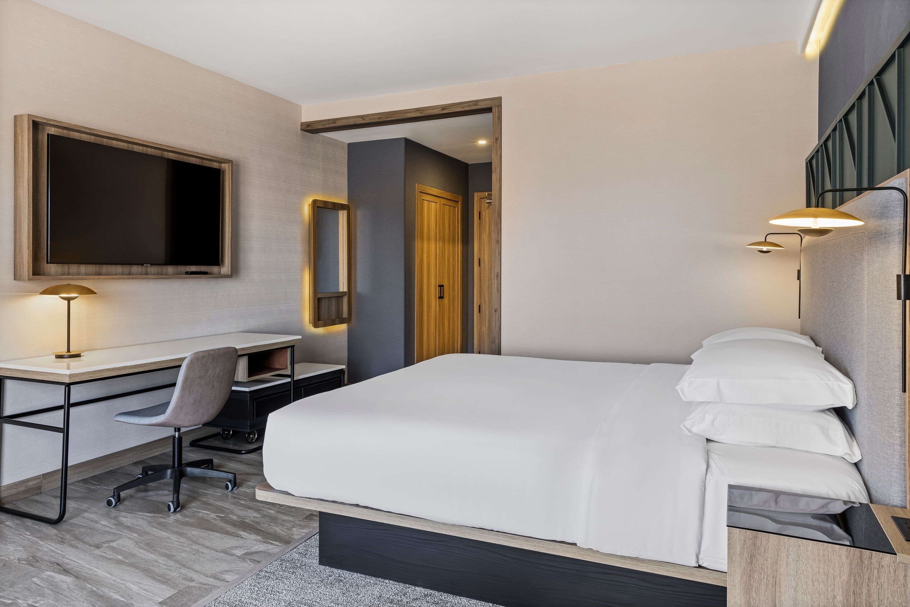 A modern hotel room with a large bed, a desk with a chair, a flat-screen TV, and a mirror.