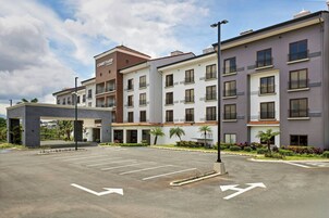 Un hotel moderno con varios pisos, estacionamiento y una zona ajardinada con palmeras.