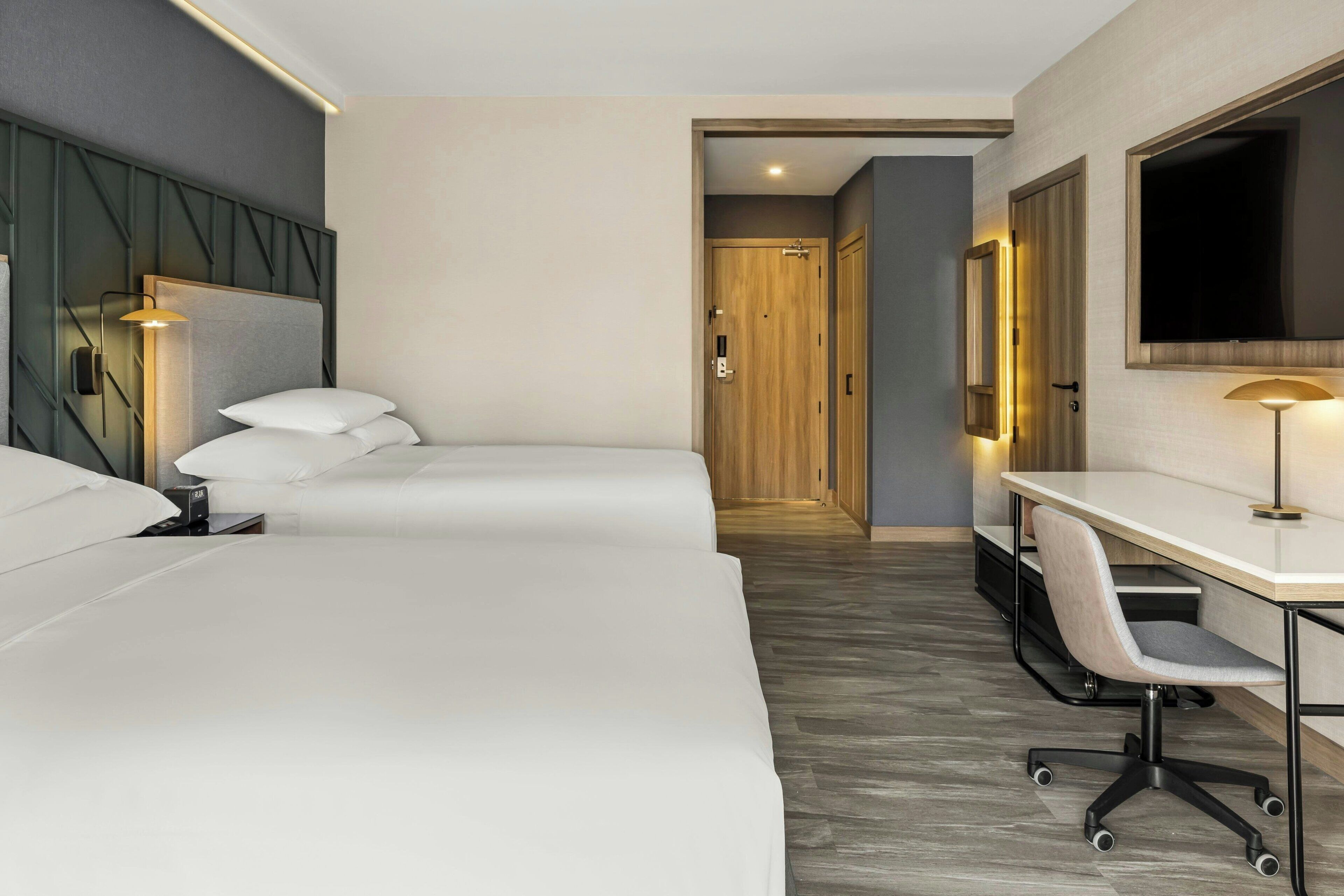 A hotel room with two beds, a desk, a chair, a TV, and a door leading to another room.