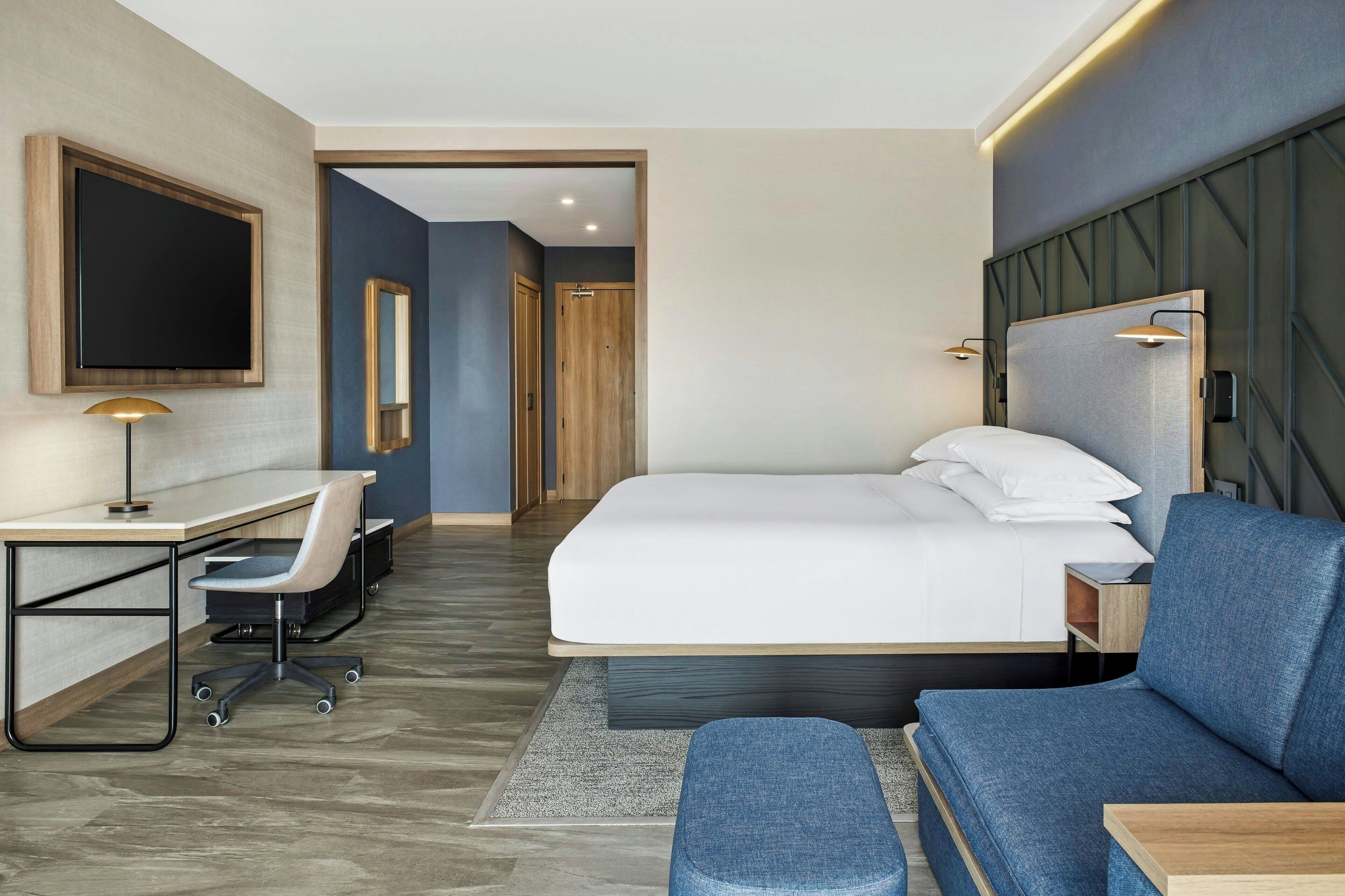 A modern hotel room with a large bed, a desk with a chair, a TV, and a seating area with a sofa and ottoman.