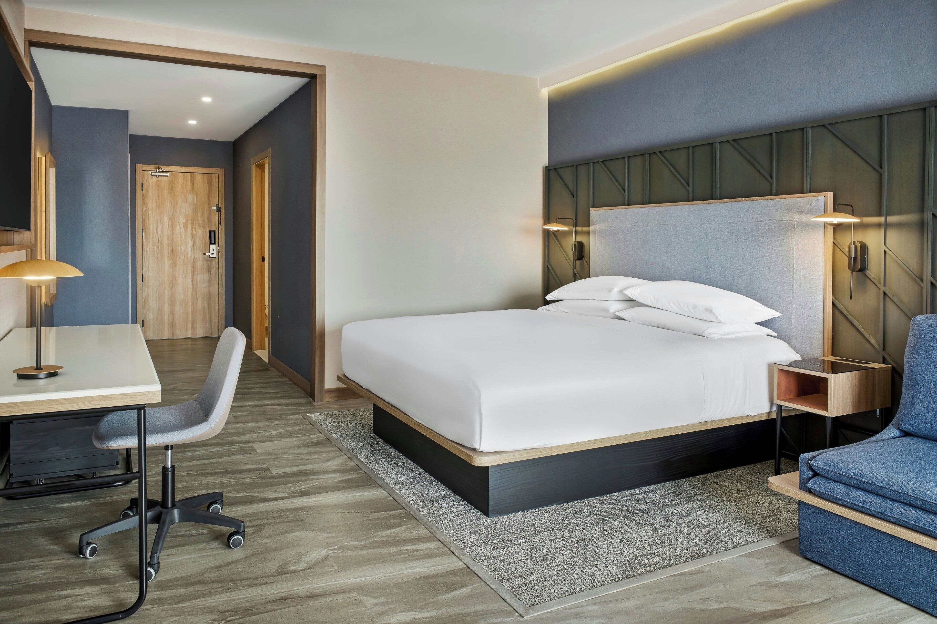 A modern hotel room with a bed, desk, chair, and a small seating area.