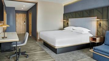 A modern hotel room with a bed, desk, chair, and a small seating area.