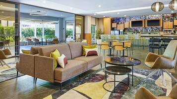 A modern hotel lobby with a sofa, chairs, a bar area, and a view of greenery outside.