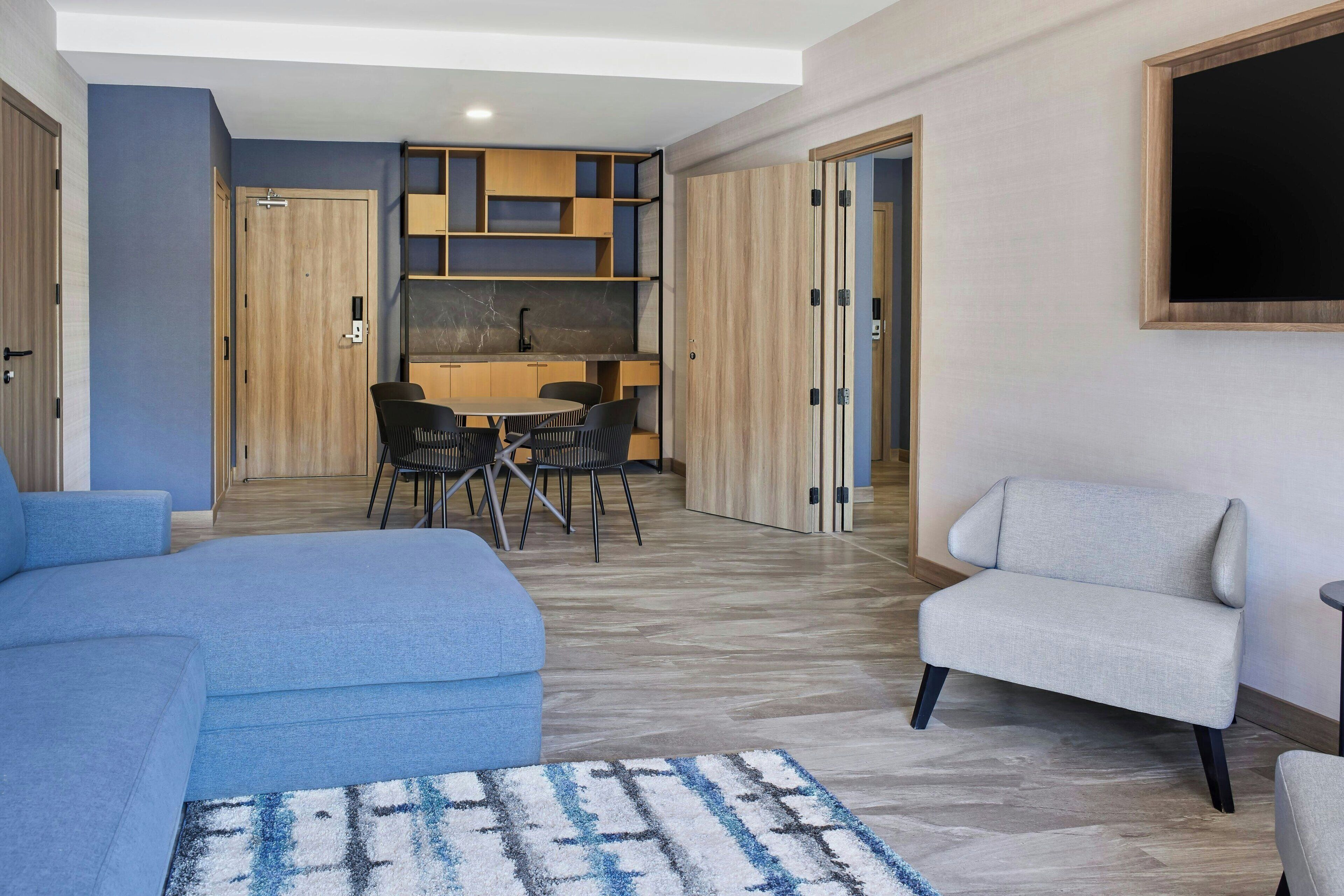 Een moderne woonkamer met een blauwe bank, een grijze fauteuil en een kamerbreed tapijt. Er is een eethoek met een tafel en stoelen, en een keuken met houten kastjes en een marmeren achterwand.