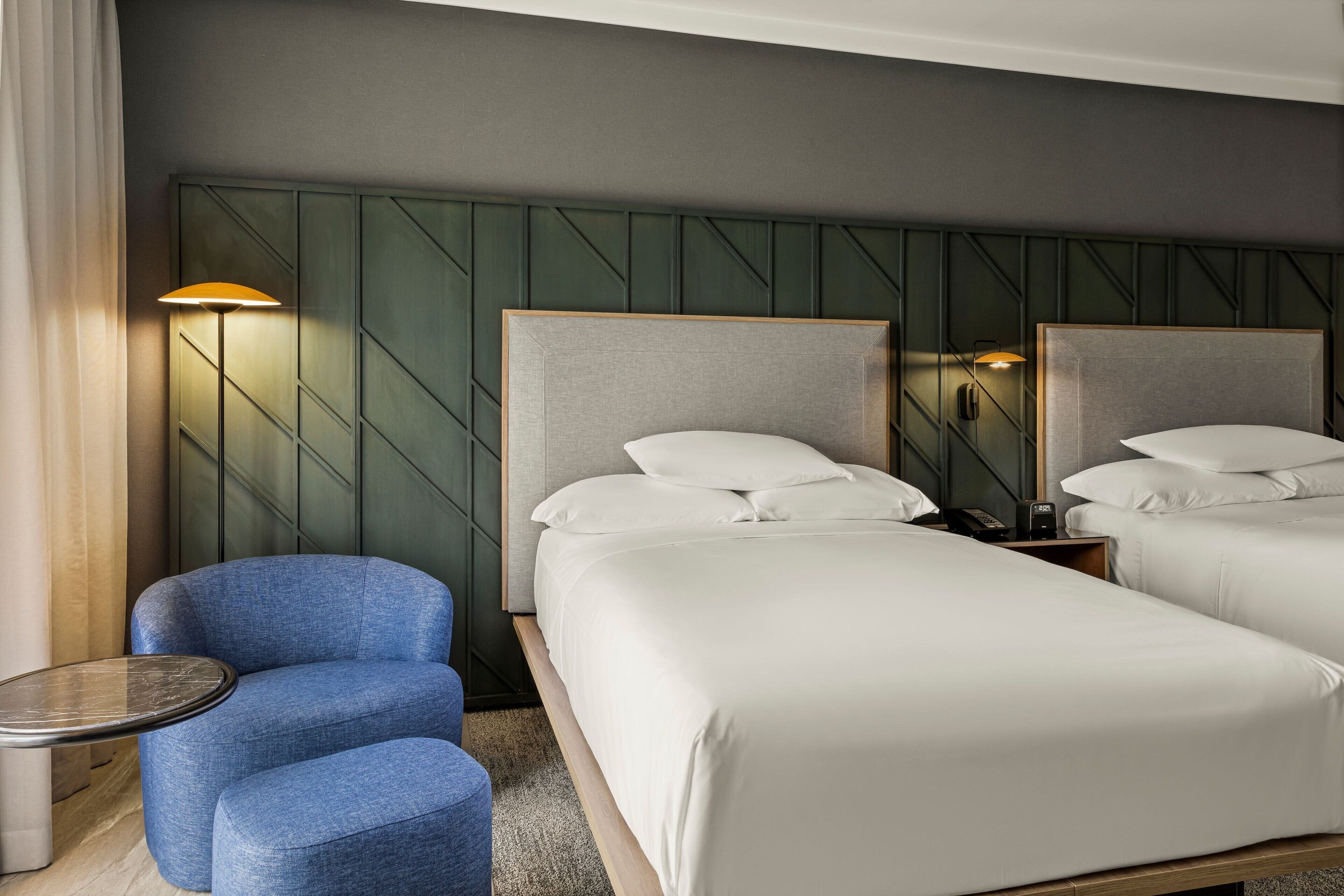 Habitació d'hotel amb dos llits, un butàc blau, una taula petita rodona i una làmpada penjada a la paret.