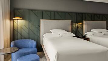 A hotel room with two beds, a blue armchair, a small round table, and a wall-mounted lamp.