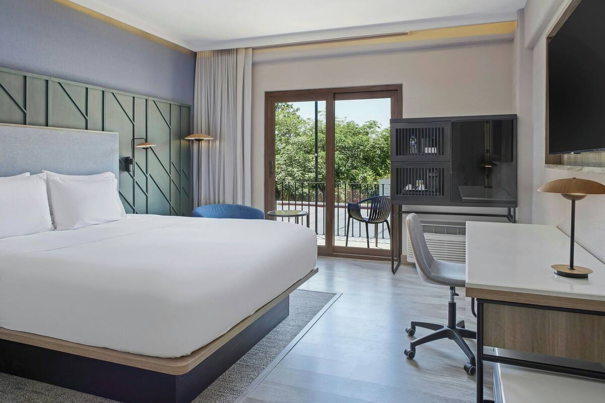 Een hotelkamer met een groot bed, een bureau met een stoel, een televisie en een balkon met uitzicht op bomen.