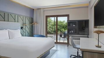 Ein Hotelzimmer mit einem großen Bett, einem Schreibtisch mit Stuhl, einem Fernseher und einem Balkon mit Blick auf Bäume.