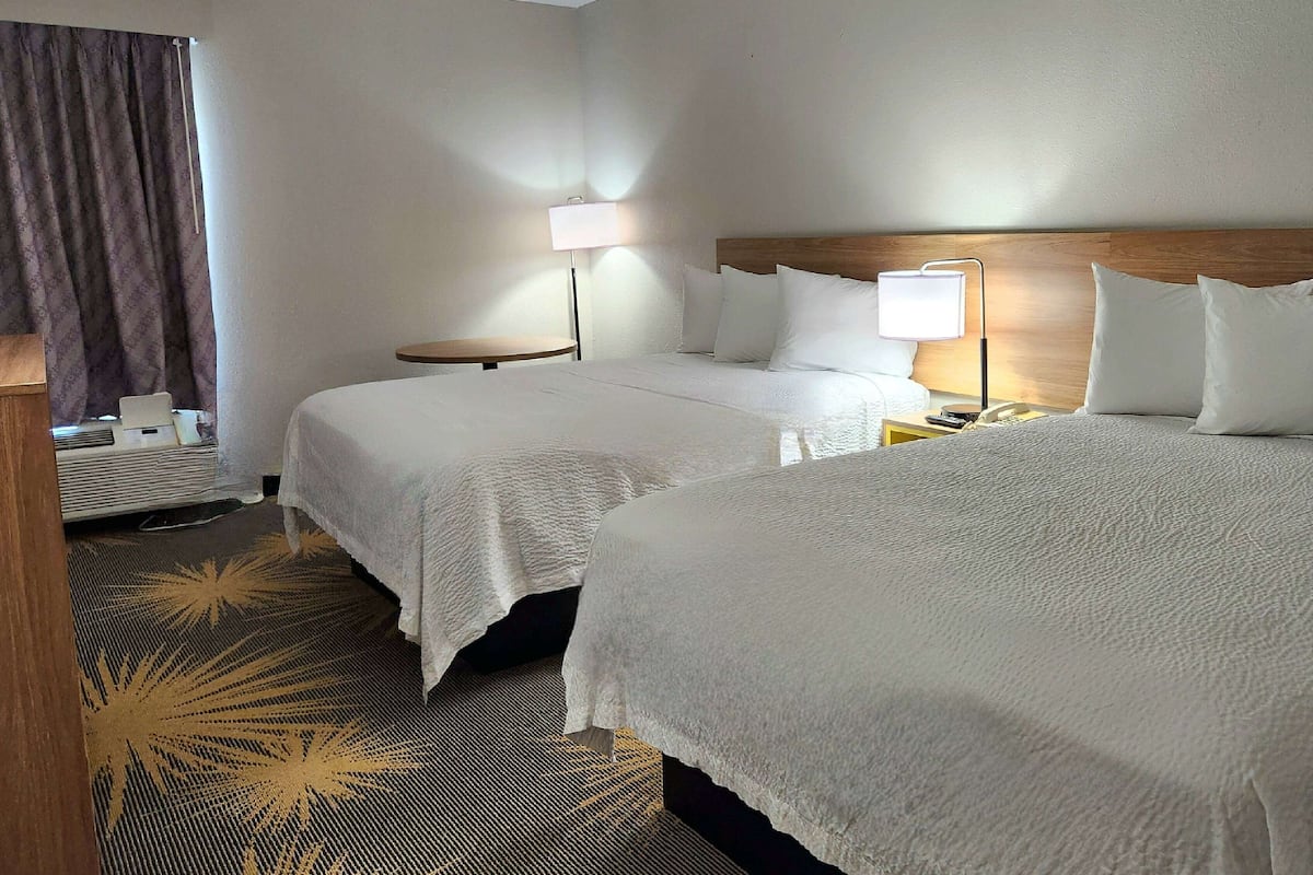 A hotel room with two beds, a bedside table with a lamp, a small round table, and a window with curtains.