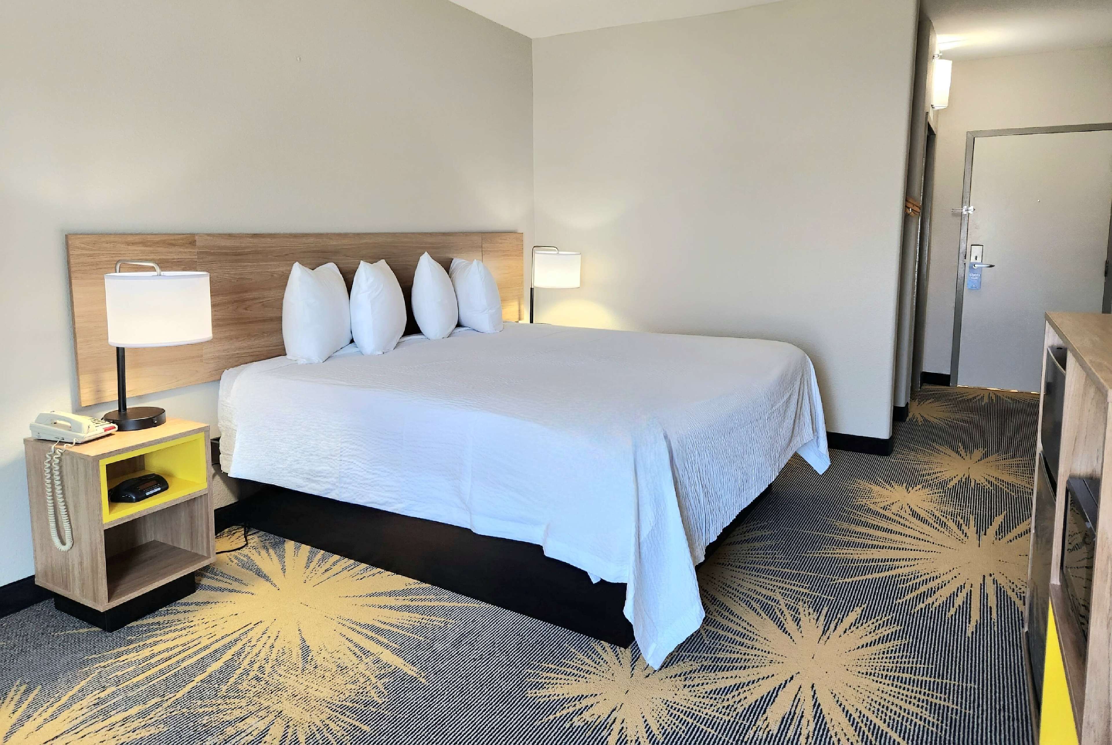A hotel room with a large bed, two bedside lamps, a wooden nightstand with a phone and a small shelf, and a patterned carpet.