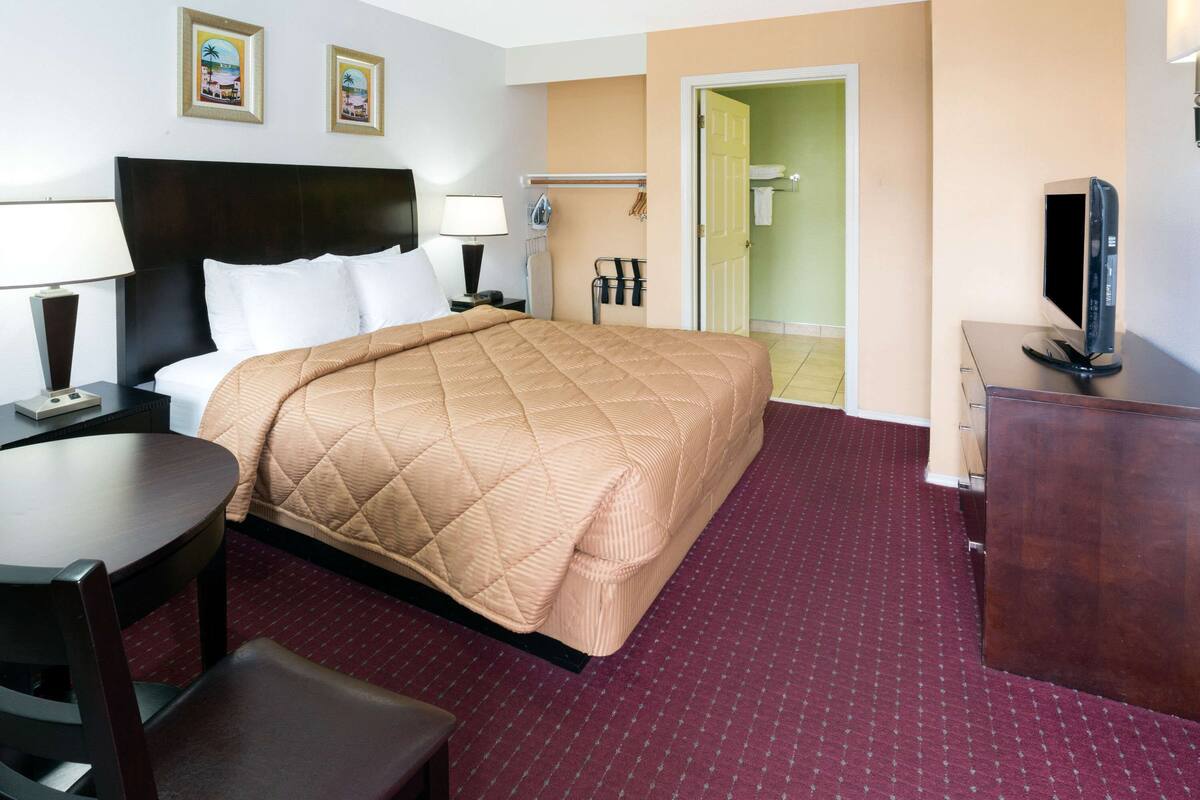 A hotel room with a bed, a desk, a chair, a TV, and a bathroom visible through an open door.