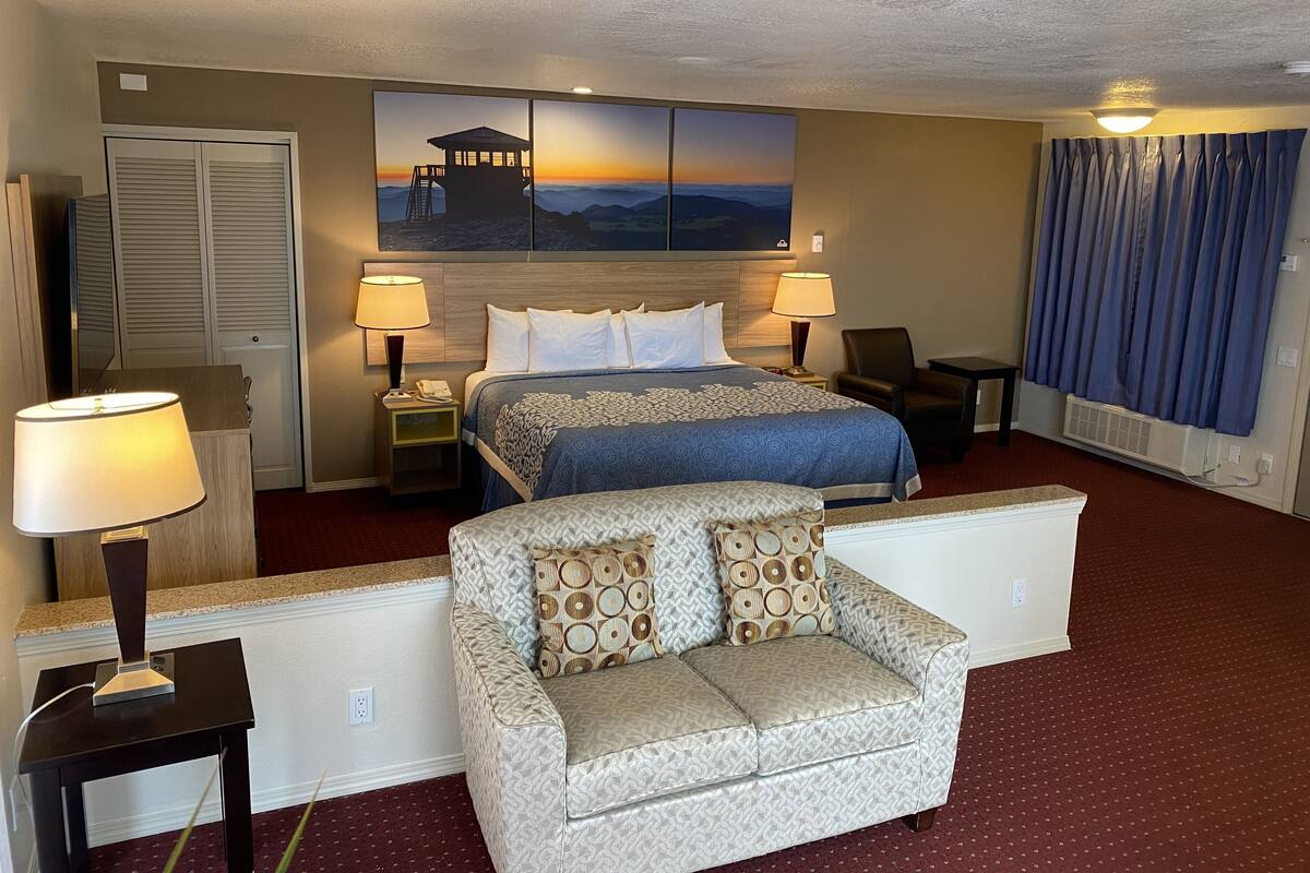 A hotel room with a king-sized bed, two bedside tables with lamps, a sofa, a large window with curtains, and a wall-mounted artwork depicting a mountain scene.