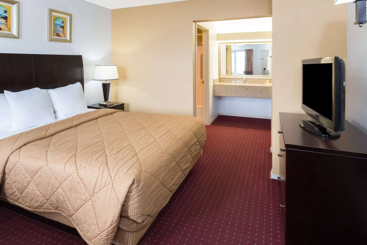A hotel room with a bed, a TV, a nightstand, a lamp, and a bathroom visible through an open door.