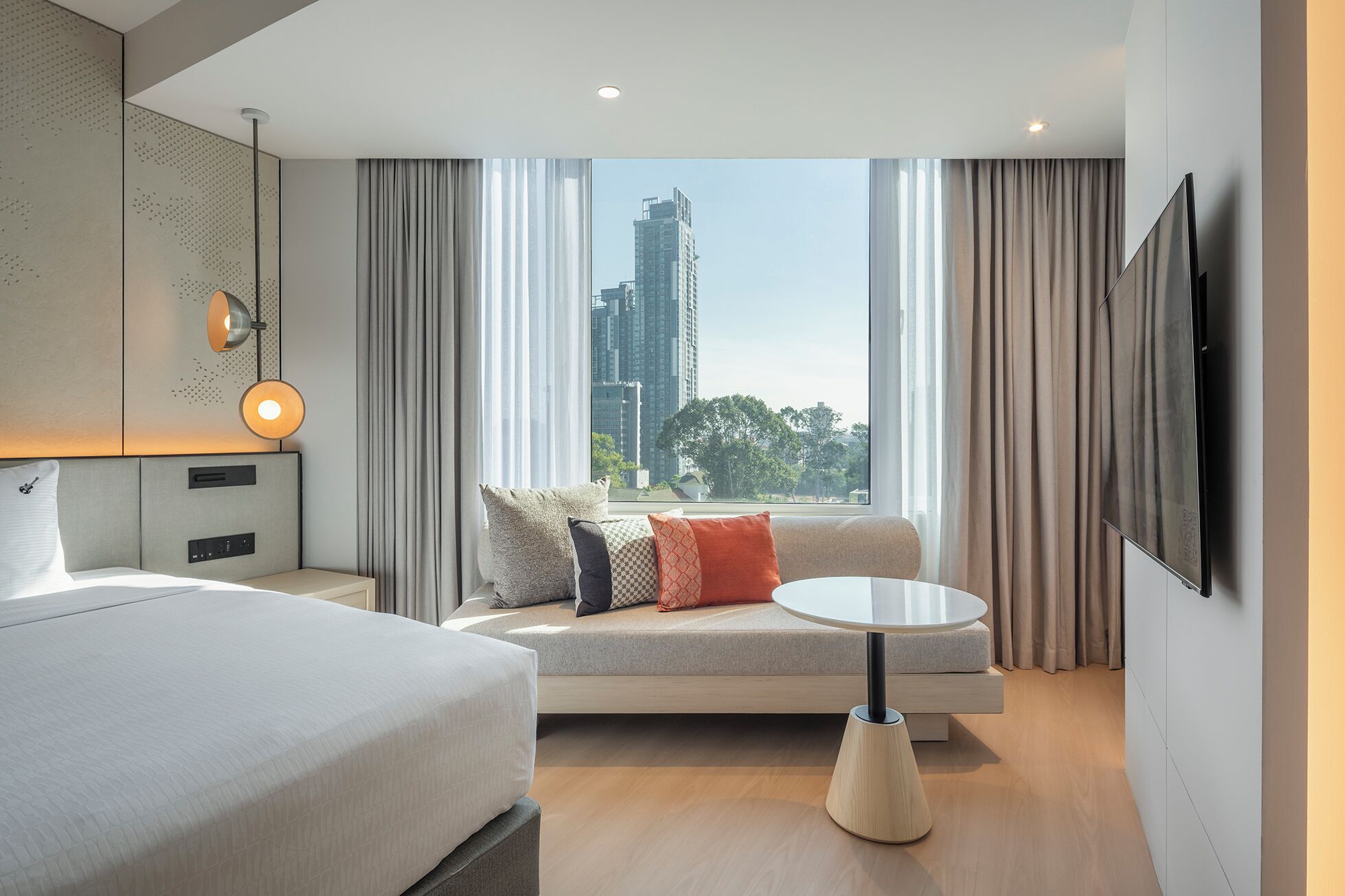 A modern hotel room with a bed, sofa, round table, and a large window with a city view.