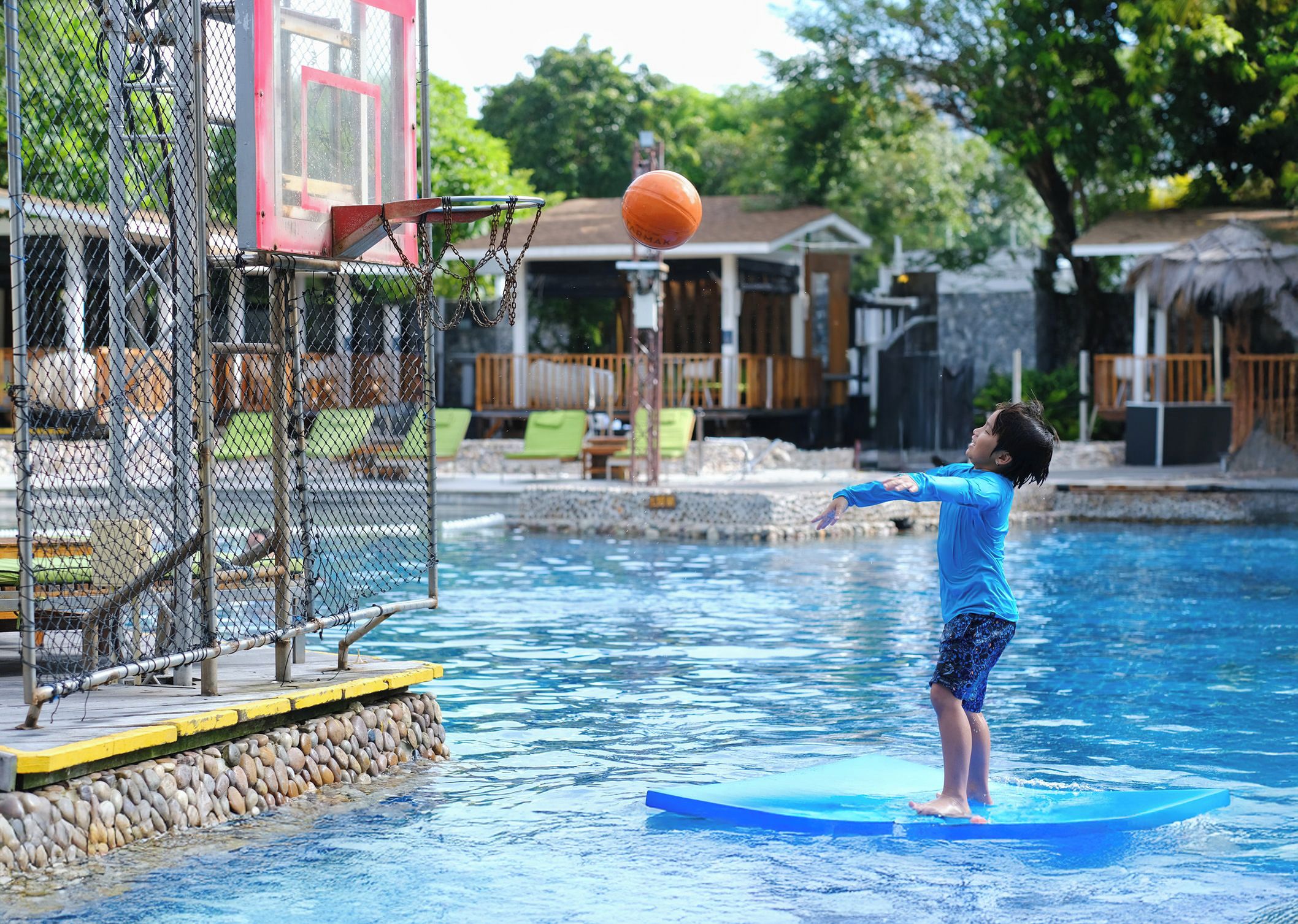 A person on a blue floating platform in a pool with a basketball hoop and net in the background.