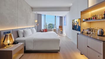 A modern hotel room with a large bed, a desk with a lamp, a view of the city, and a minibar.