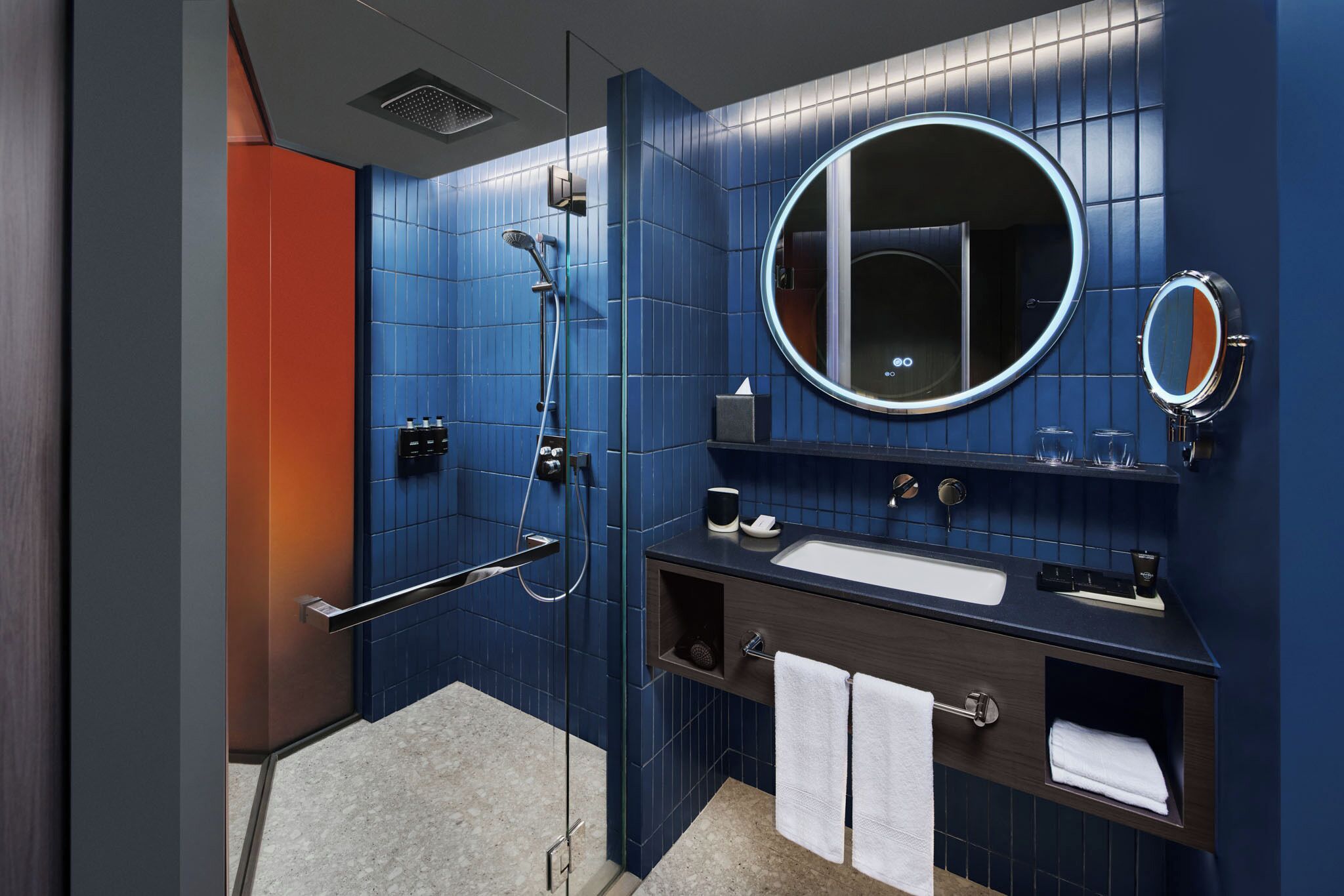 A modern bathroom with blue tiles, a round mirror, a sink, and a shower.