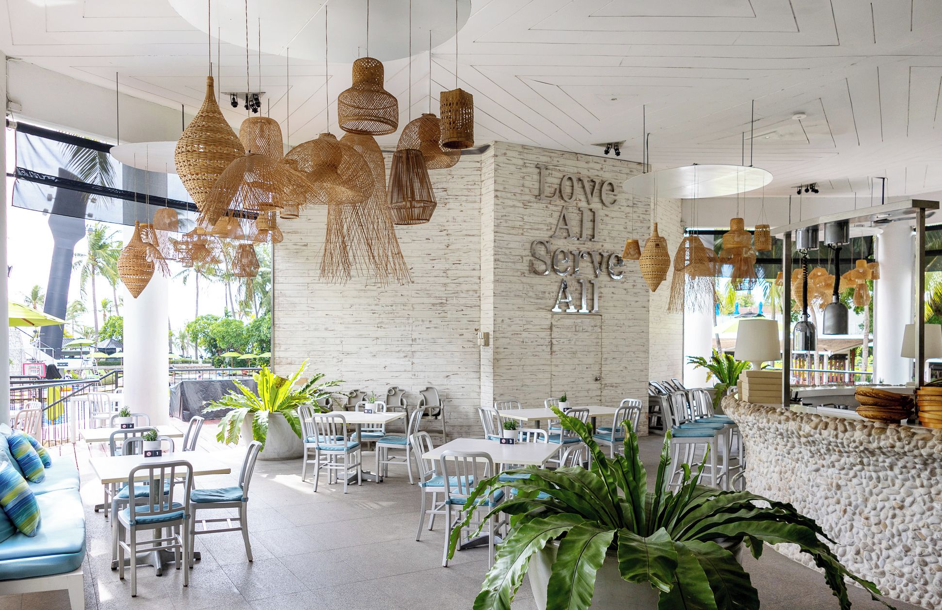 Un interior moderno con lámparas de mimbre colgantes, una pared de ladrillo blanco con el texto 'Love All Servants' y una zona de estar con mesas y sillas blancas.