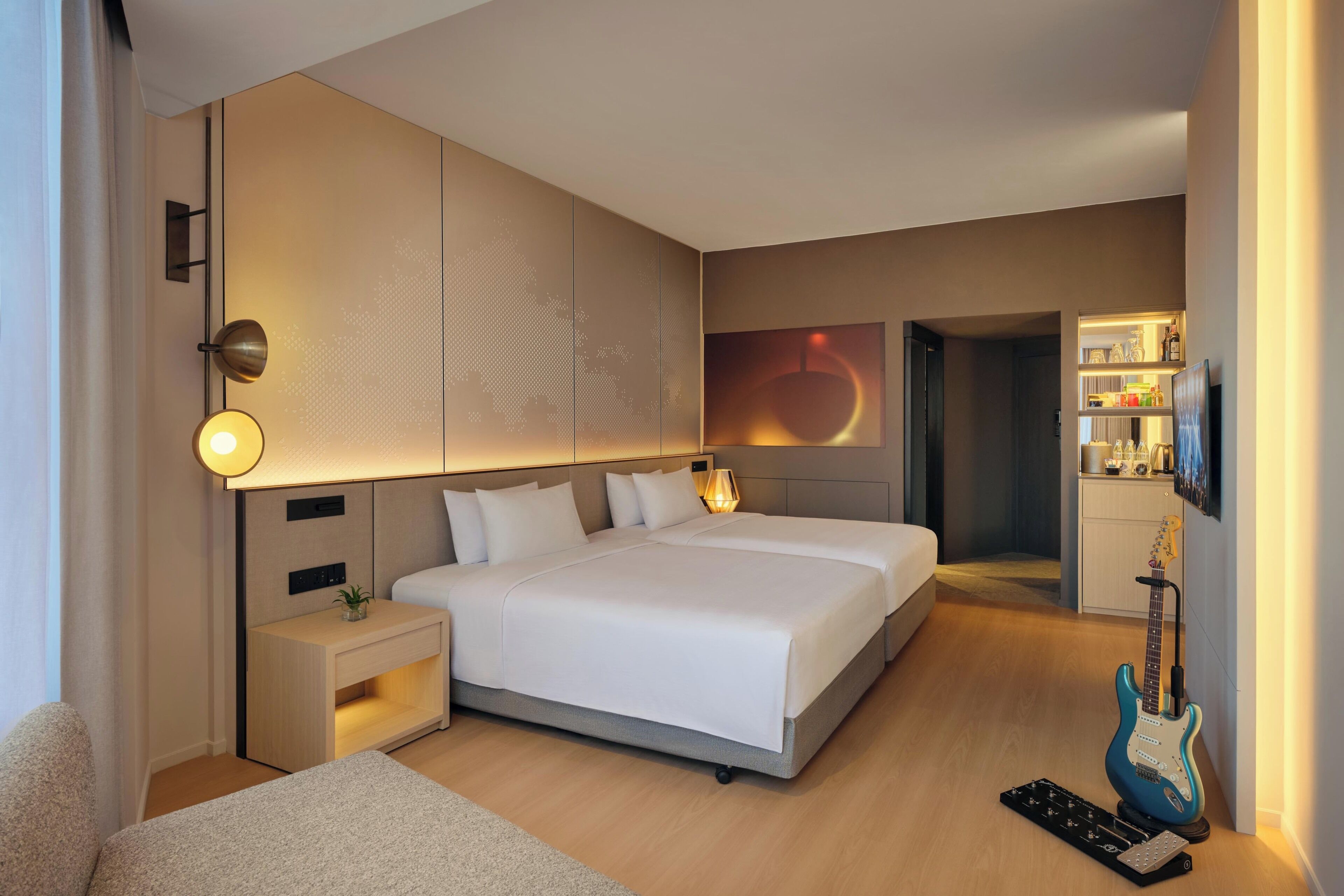 A modern hotel room with a large bed, bedside tables, a desk, a guitar, and a TV.