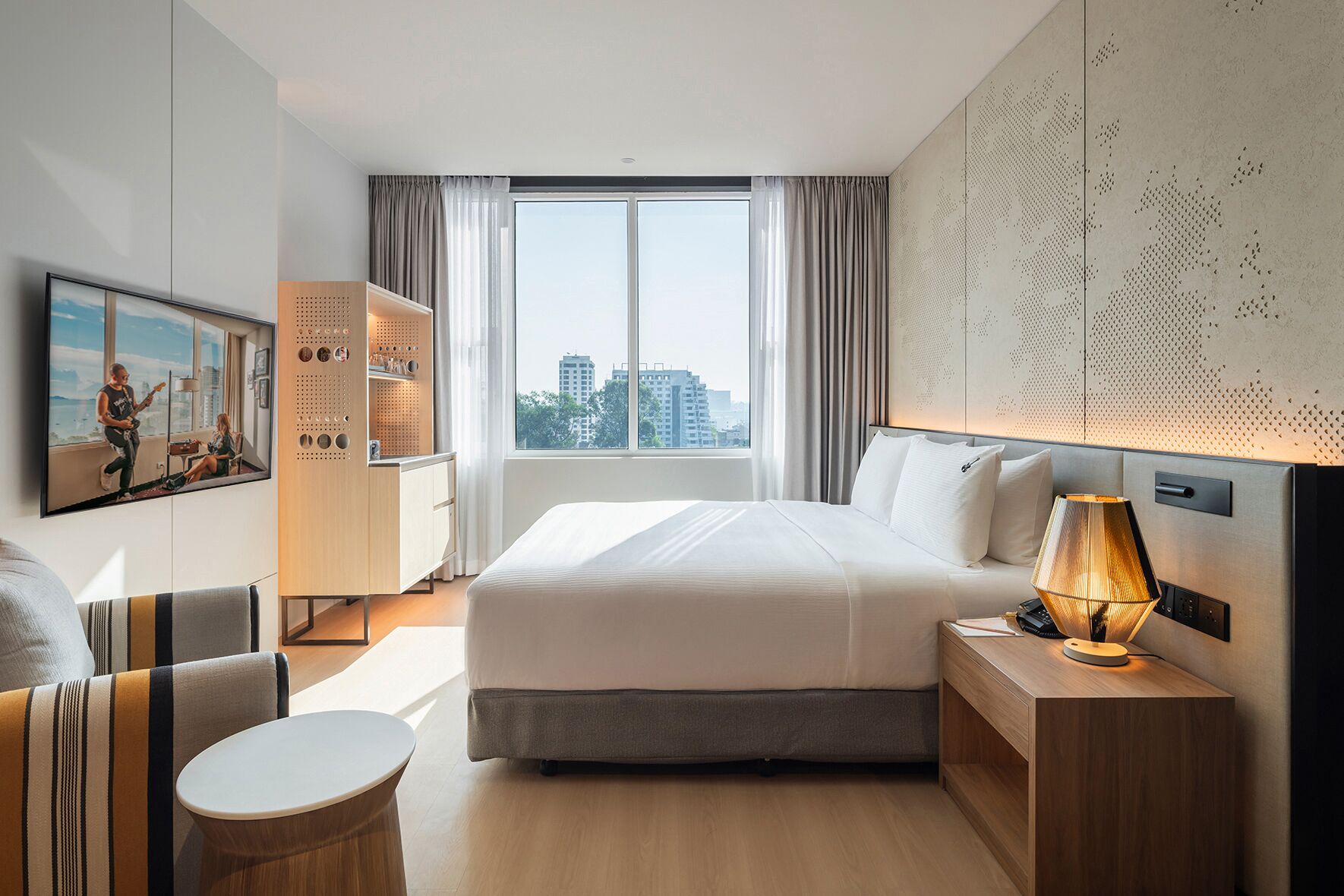 A modern hotel room with a large bed, a desk with a lamp, a small round table, and a city view through the window.