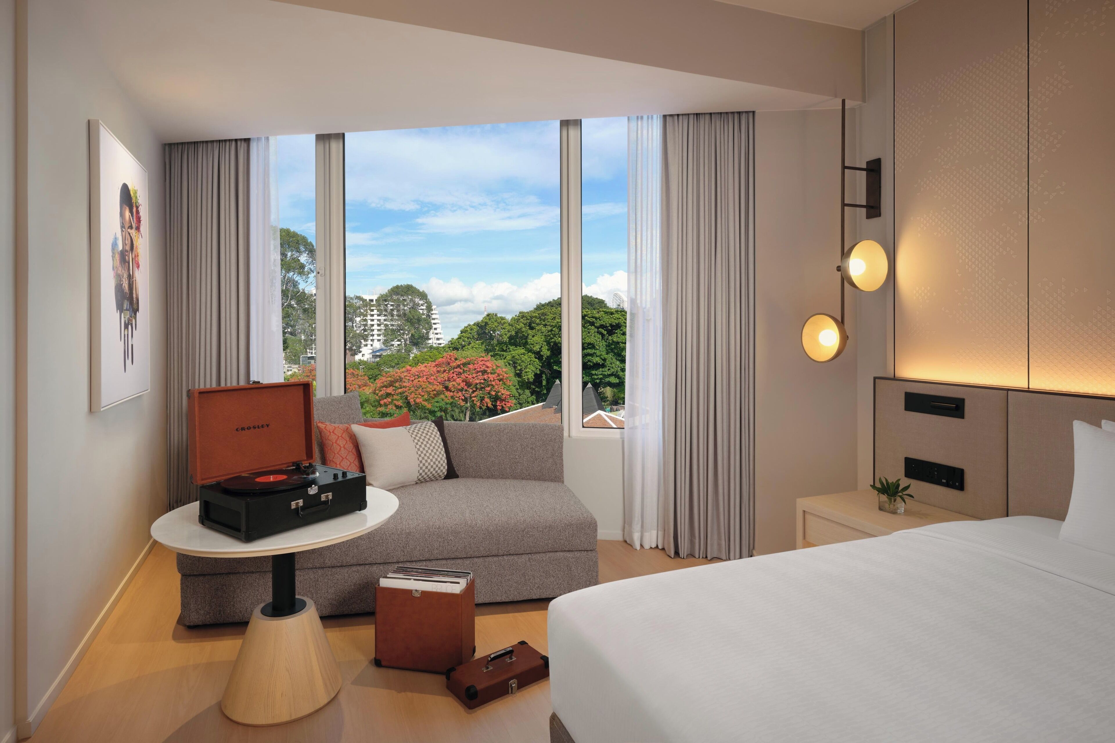 A modern hotel room with a bed, a seating area with a table, a small side table, and a large window with curtains.