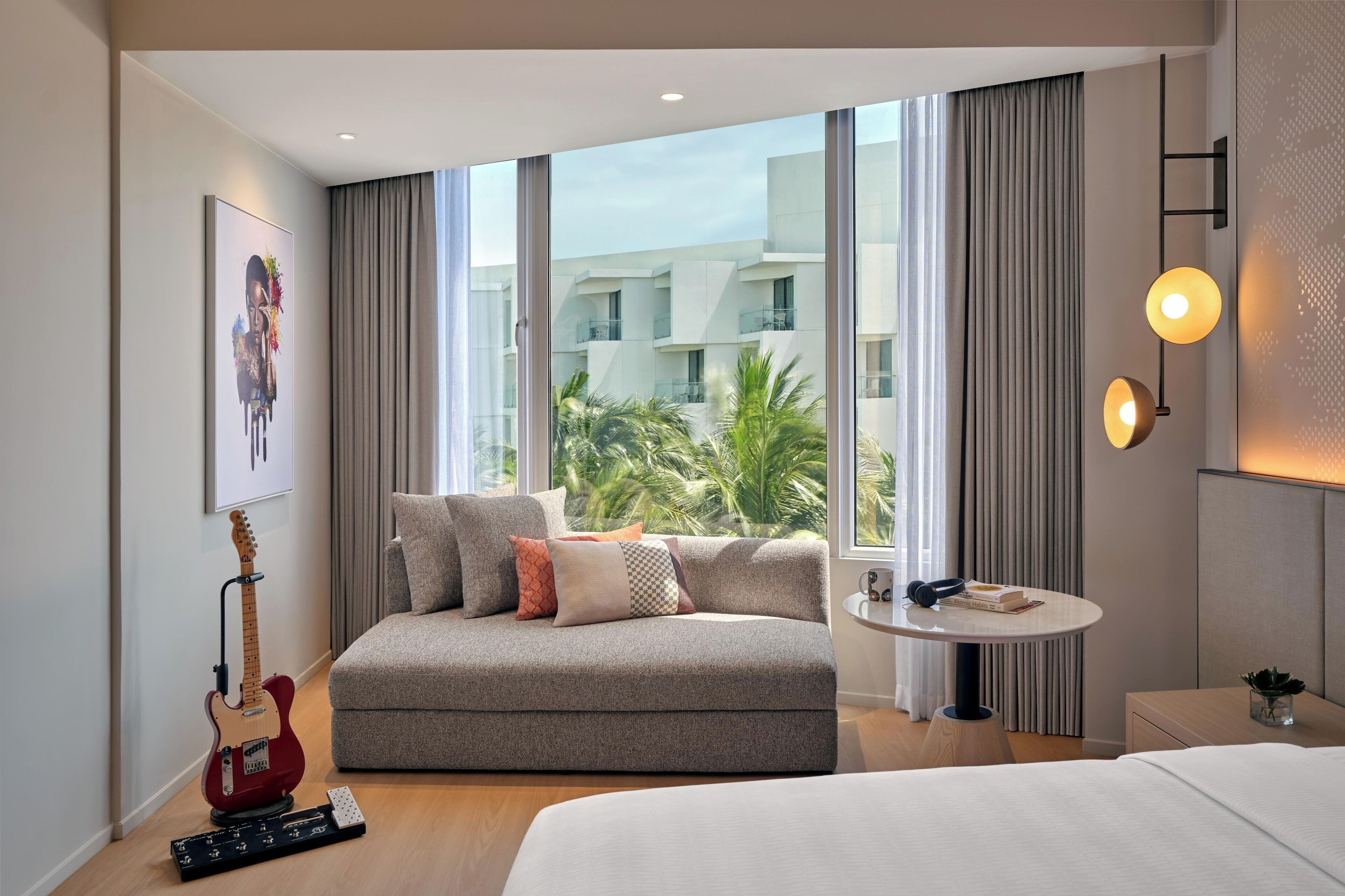 A modern hotel room with a sofa, a small table, a guitar, and a view of palm trees through large windows.