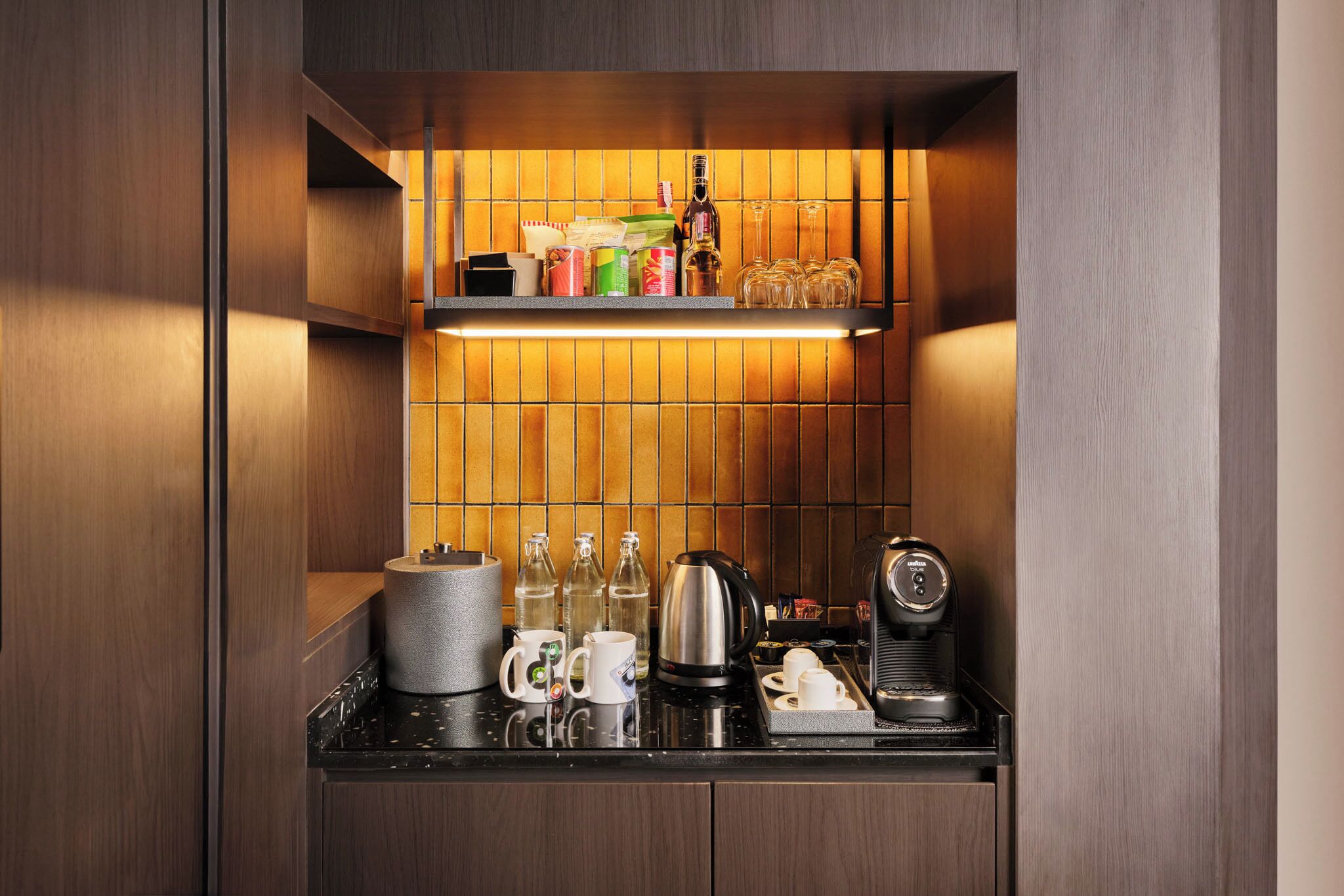 A compact kitchen with a coffee machine, a kettle, and a shelf with various items.