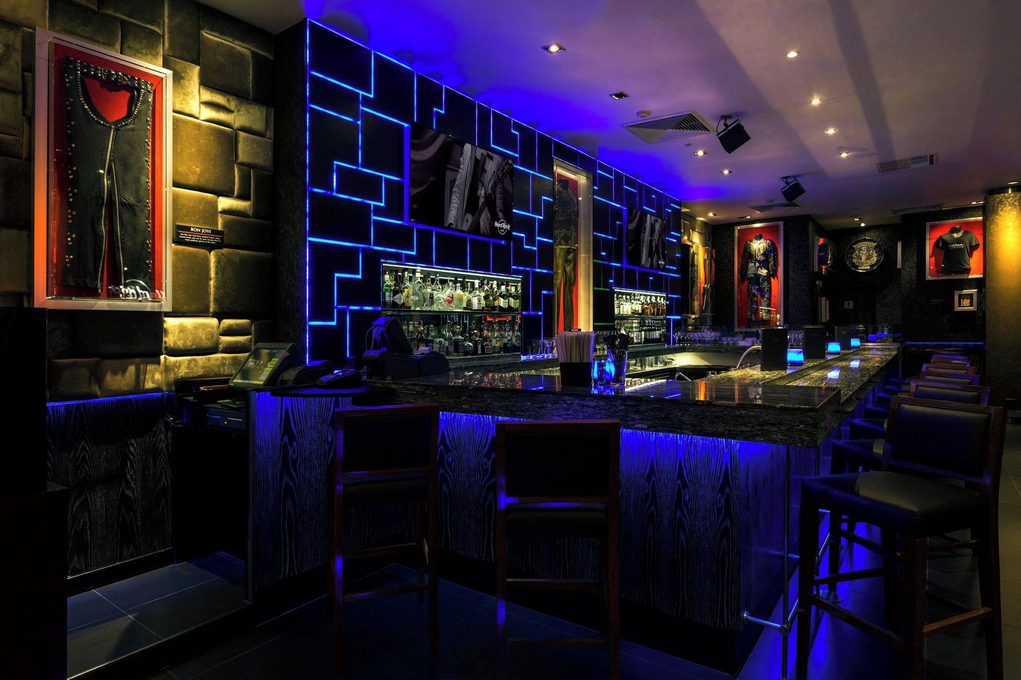 Uma área de bar com balcão, banquetas e uma parede com iluminação geométrica.