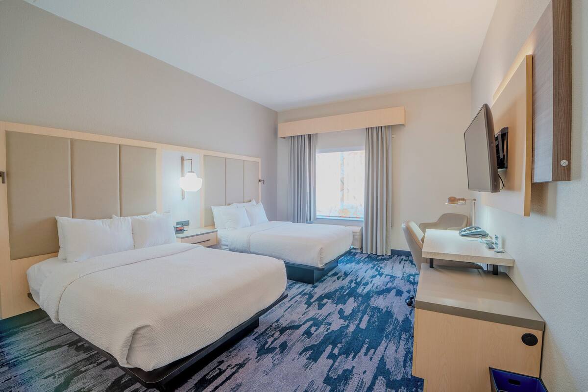 A hotel room with two beds, a desk, a chair, a TV, and a window with curtains.