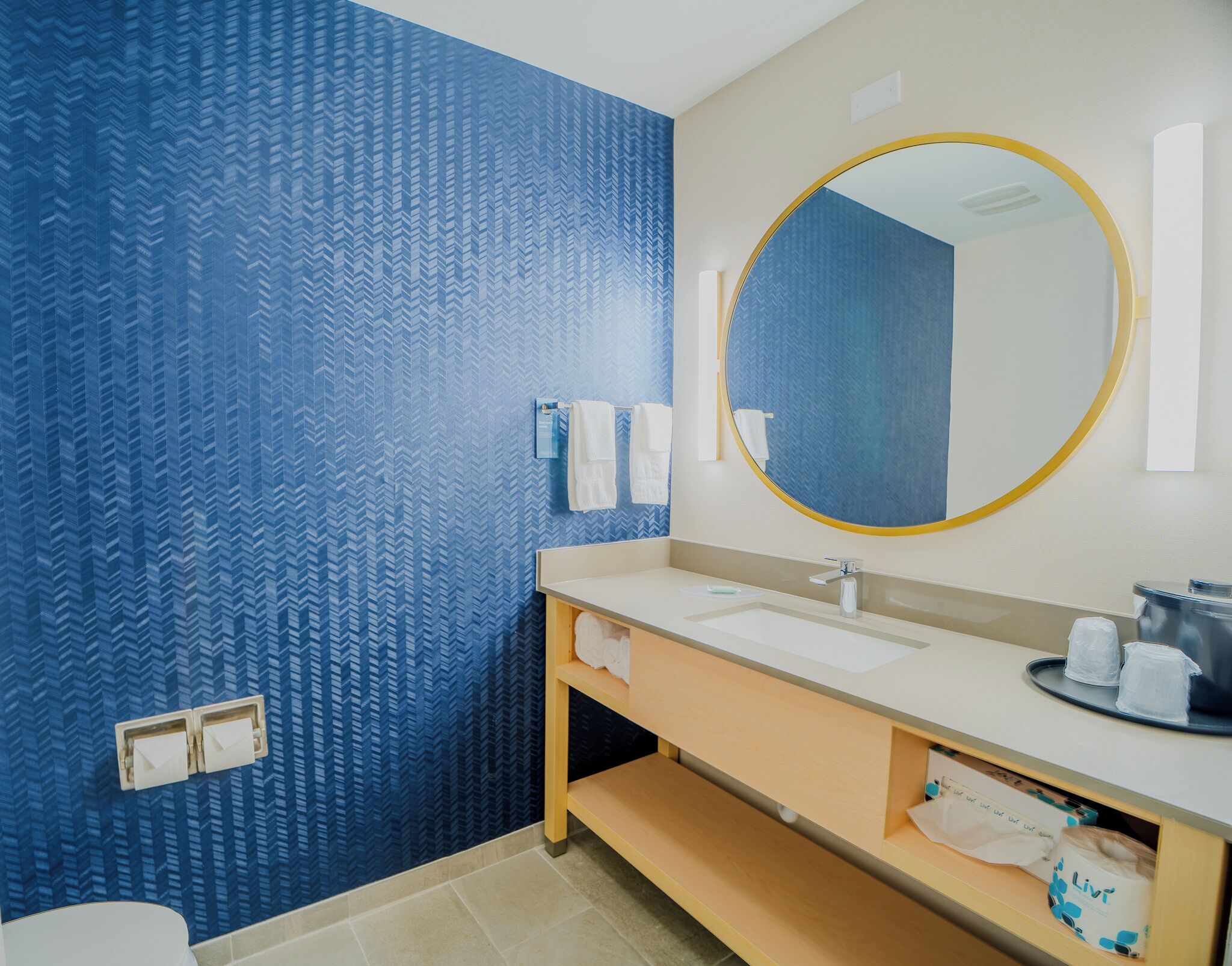 A bathroom with a blue patterned wall, a round mirror with a gold frame, a white sink, and a wooden vanity.