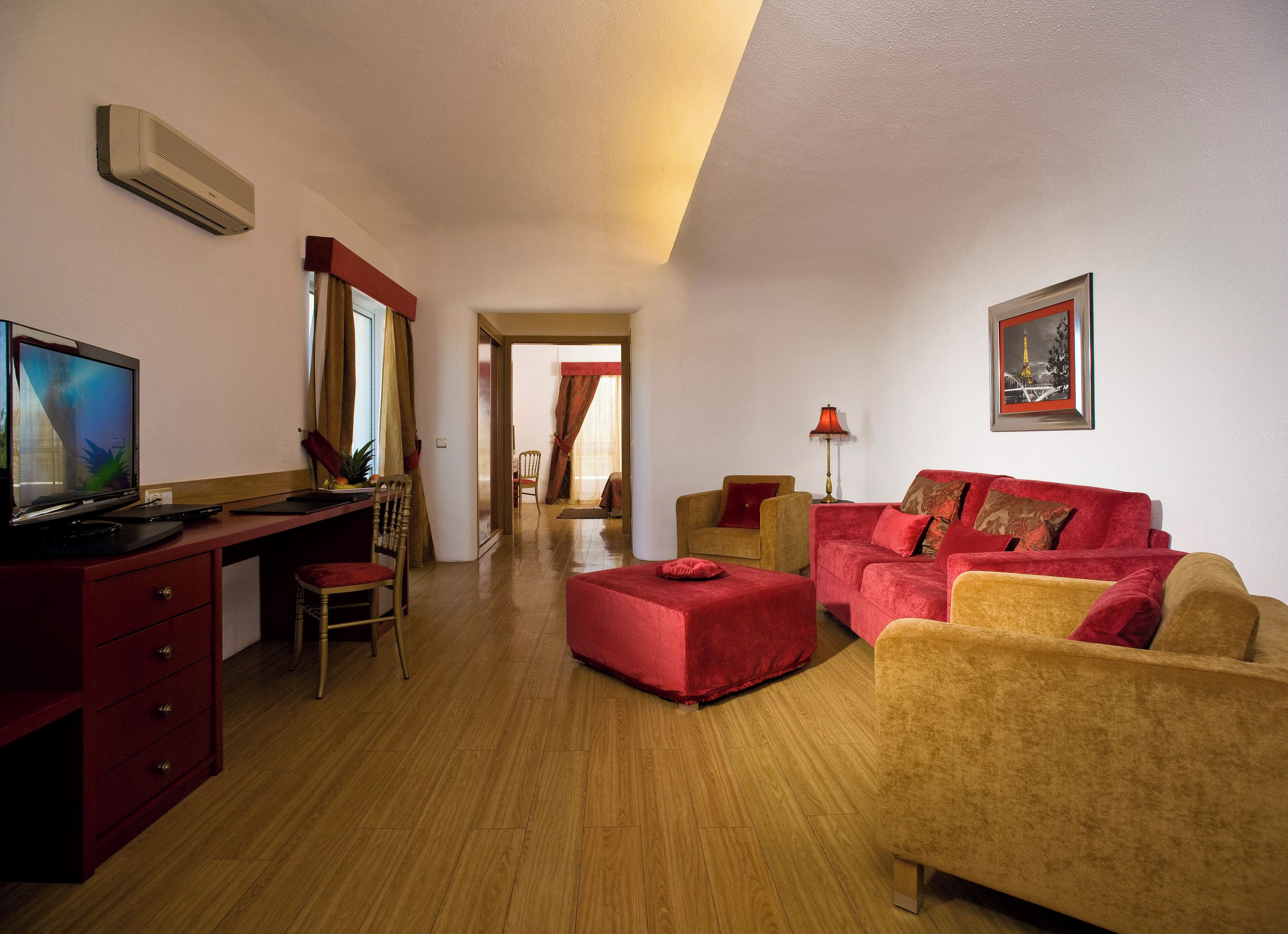 A room with a desk, computer, chair, sofa, and ottoman. There is a lamp and a picture on the wall.