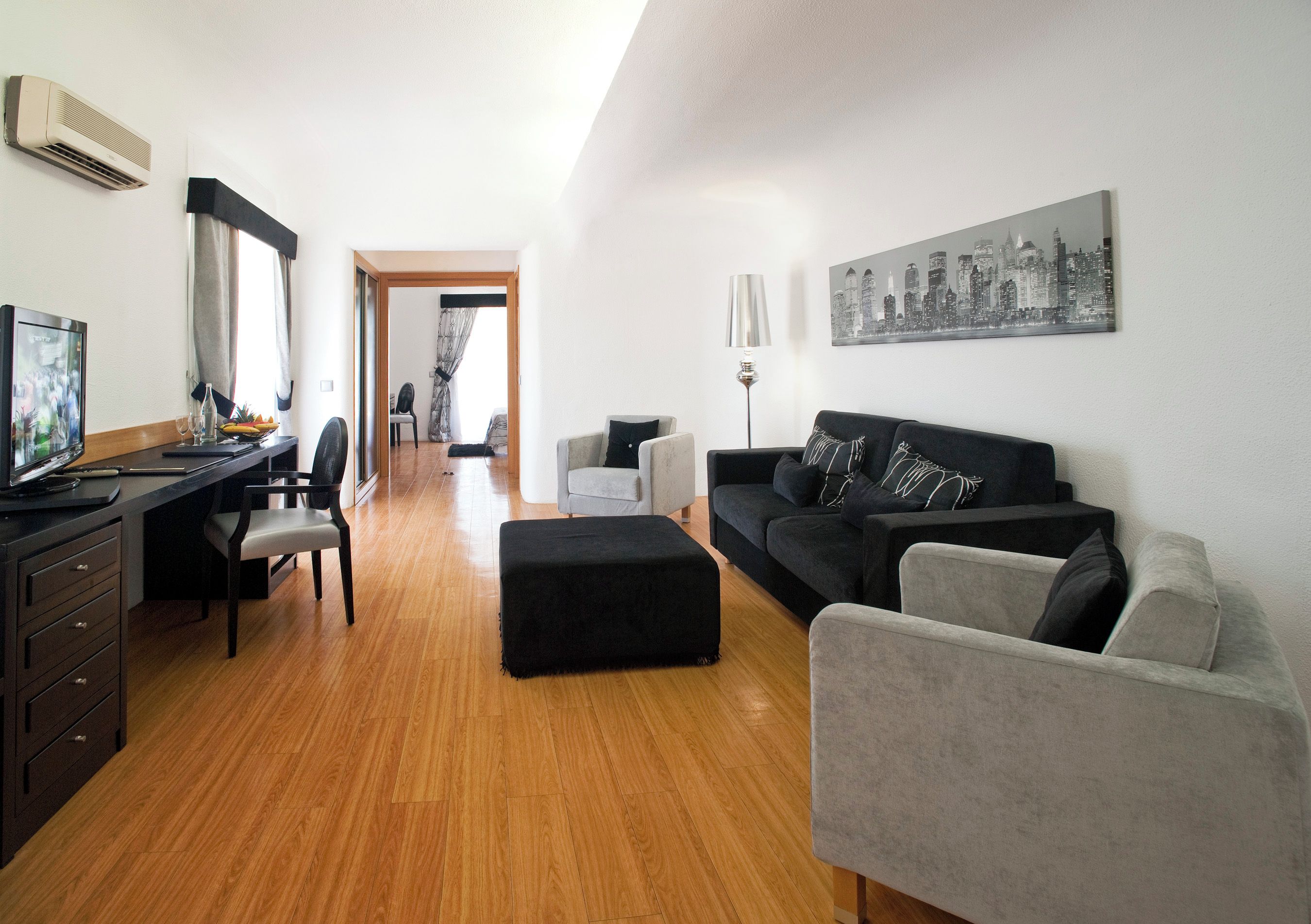 A modern living room with a wooden floor, a black sofa, a grey armchair, a black ottoman, a desk with a computer, and a cityscape painting on the wall.
