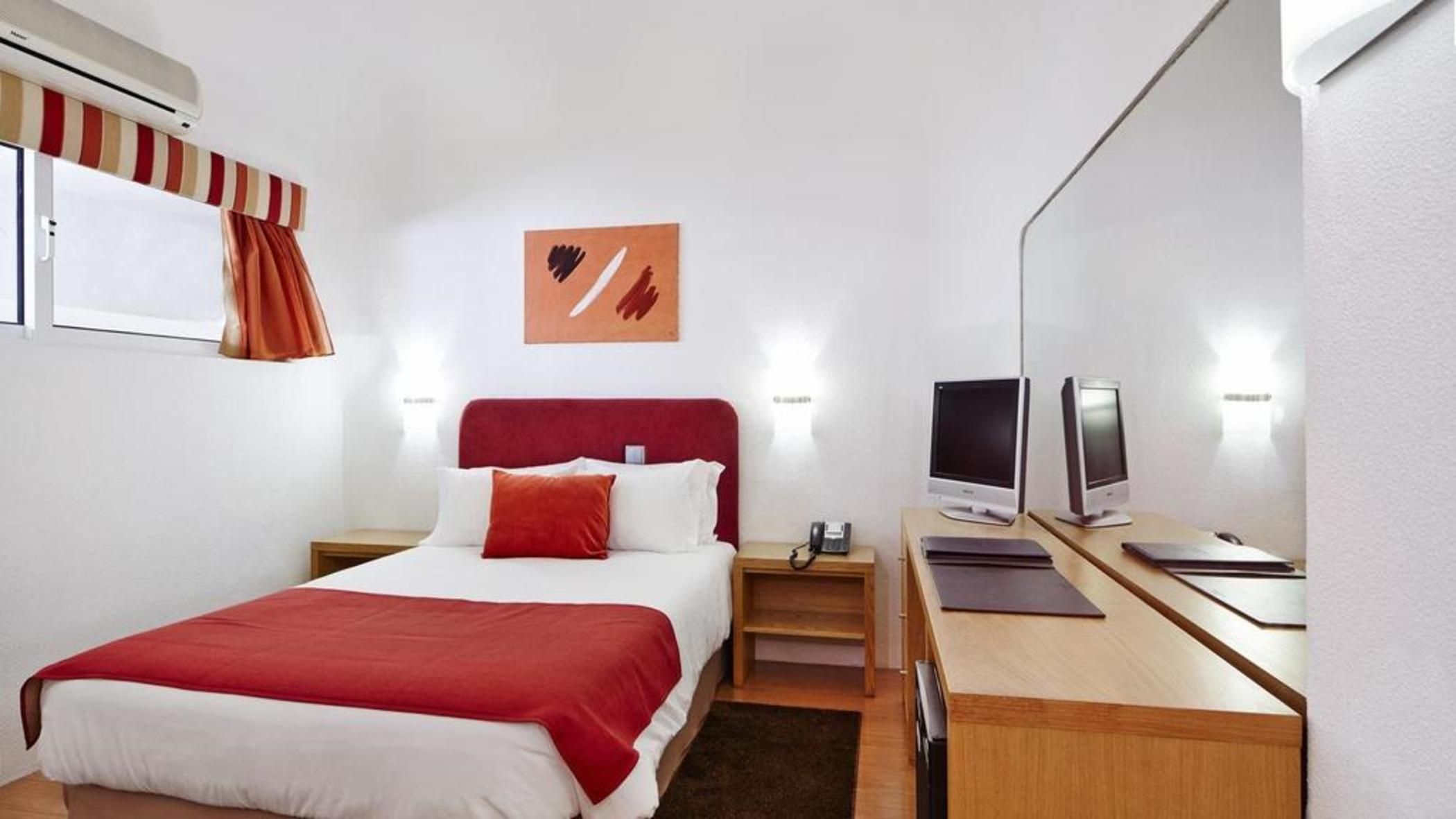 A hotel room with a bed, bedside table, TV, and a desk with a chair.