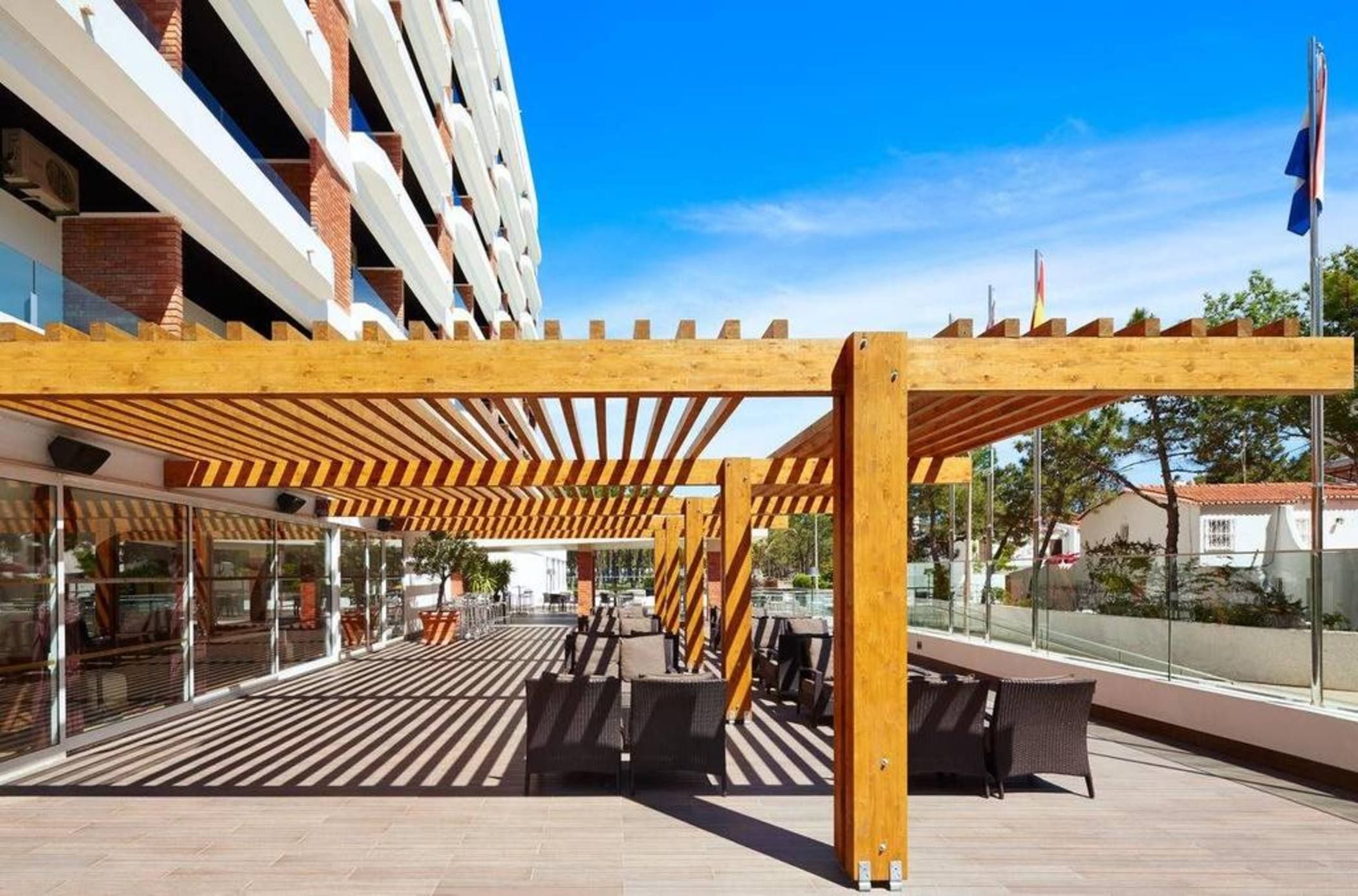 Een terras met een houten pergola, tuinmeubilair en uitzicht op een gebouw met een glazen gevel.