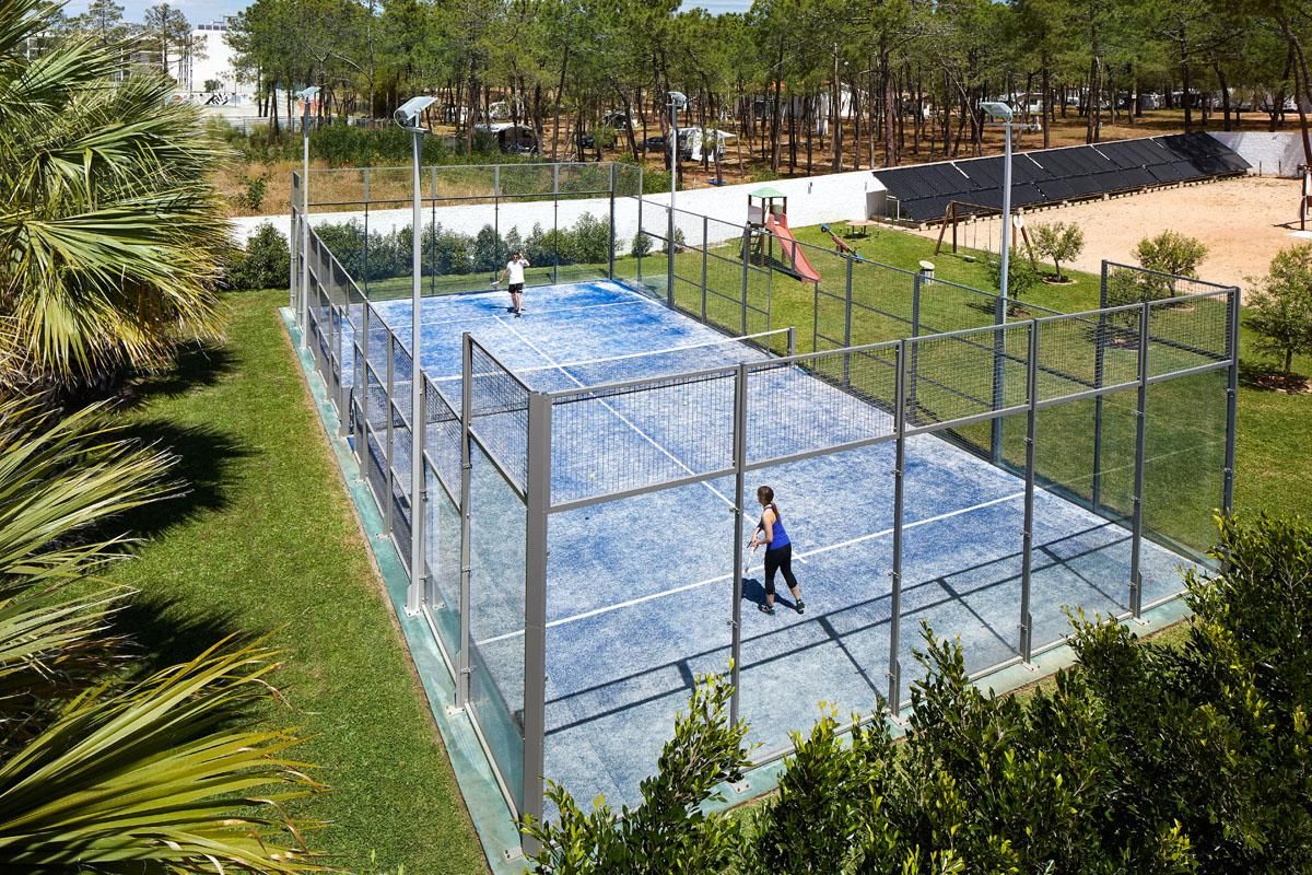 Una cancha de tenis rodeada de vegetación y un parque infantil, delimitada por una valla metálica.
