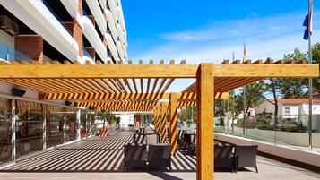 Un espace extérieur aménagé avec des pergolas et des chaises en bois, offrant une vue sur un bâtiment moderne doté de balustrades en verre.