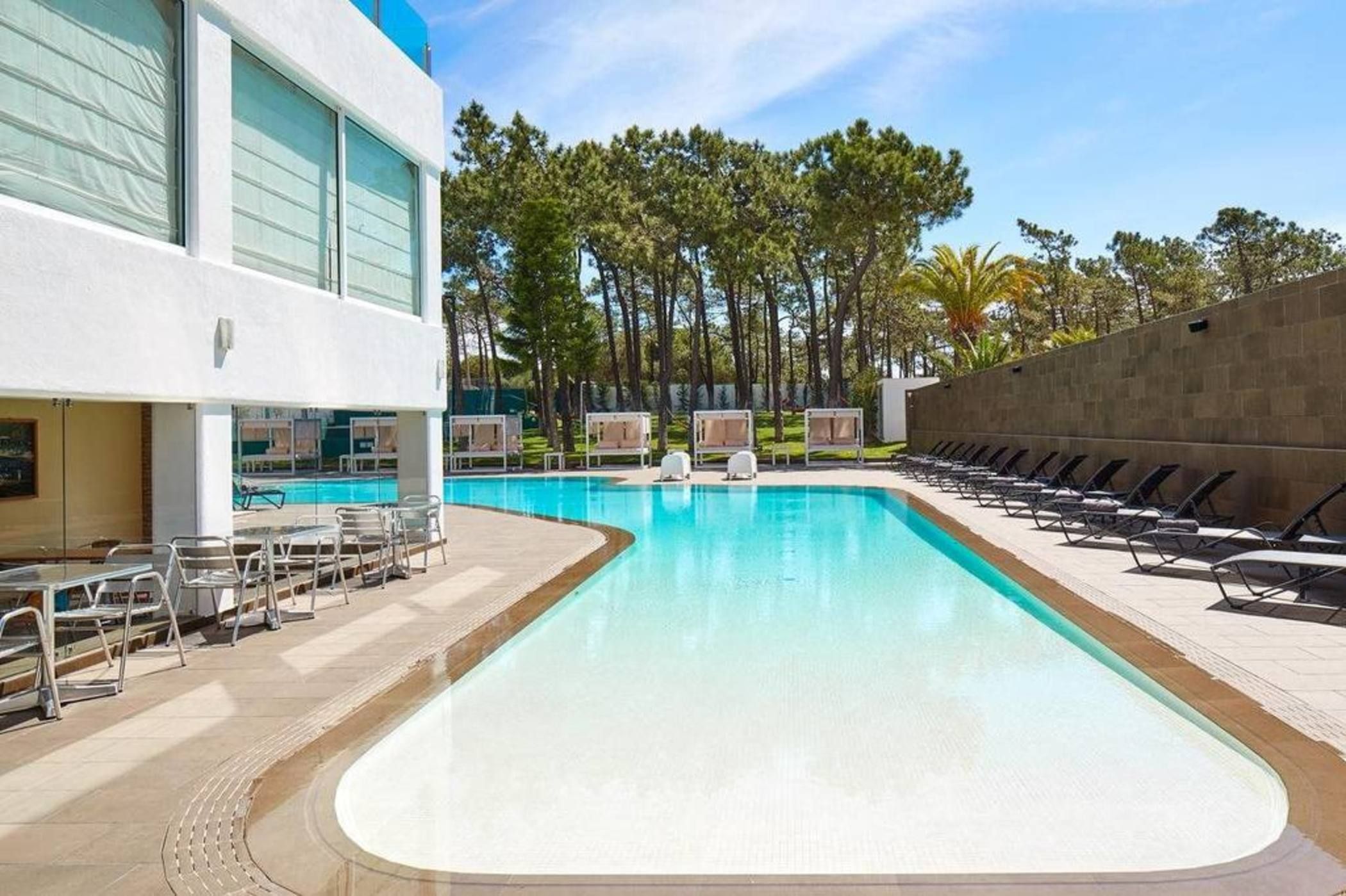 A swimming pool with lounge chairs and sunbeds, surrounded by trees and a building with large windows.