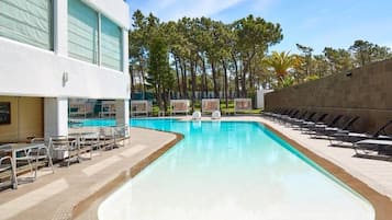 A swimming pool with lounge chairs and sunbeds, surrounded by trees and a building with large windows.