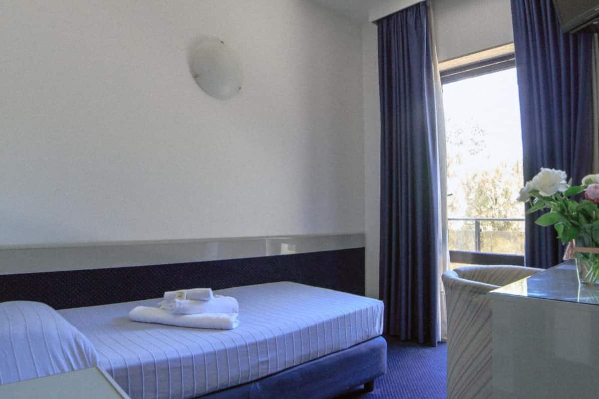Une chambre d’hôtel avec un lit, un bureau, une chaise, une fenêtre avec des rideaux et un vase de fleurs.