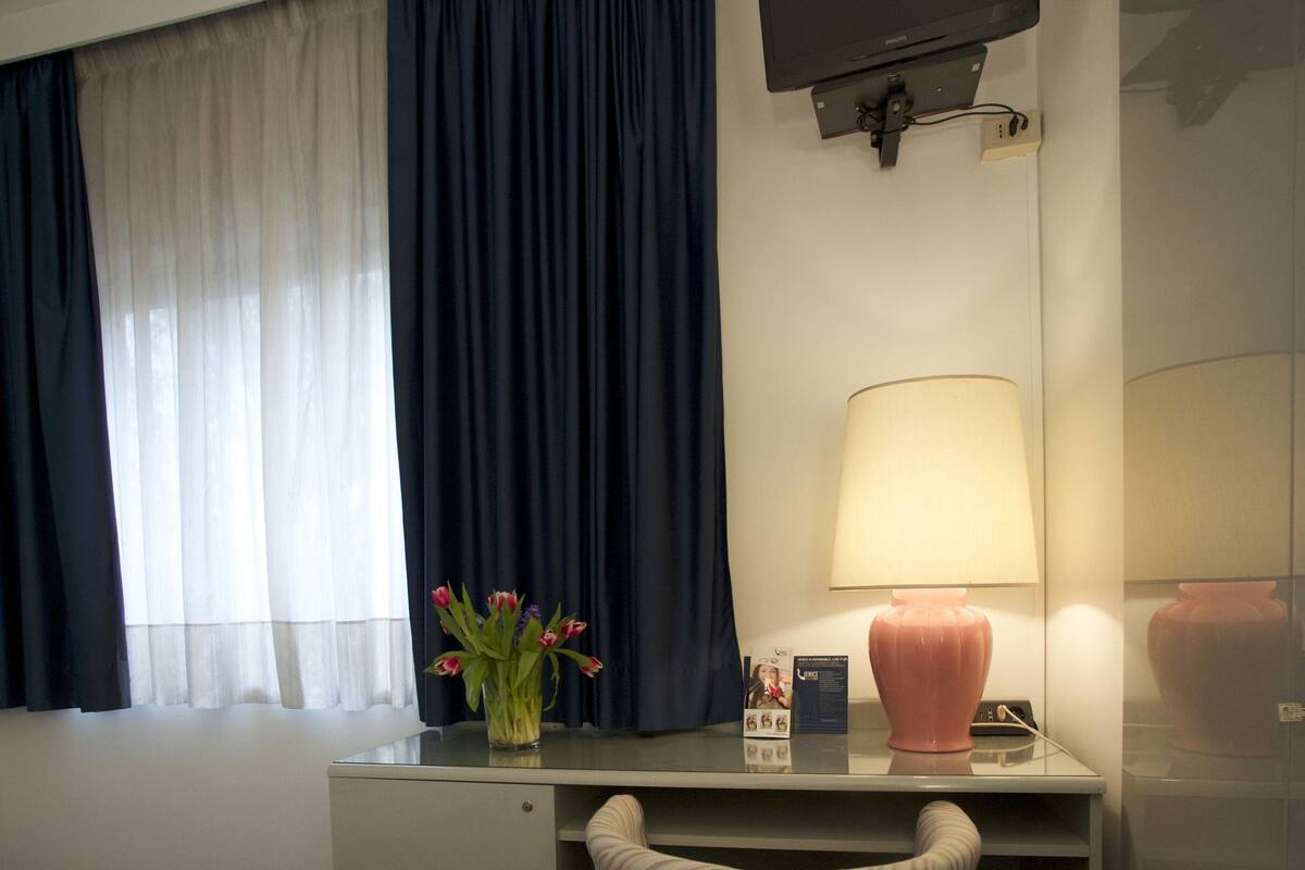 Une chambre d’hôtel comprenant un lit, un bureau, une chaise, un téléviseur fixé au mur, une lampe et une fenêtre avec des rideaux.