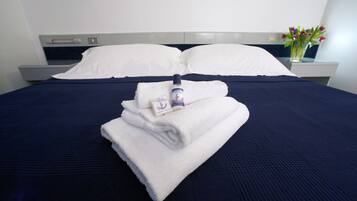 A neatly made bed with white pillows and a folded towel, a bottle of Club Hotel toiletries, and a vase with flowers on a bedside table.