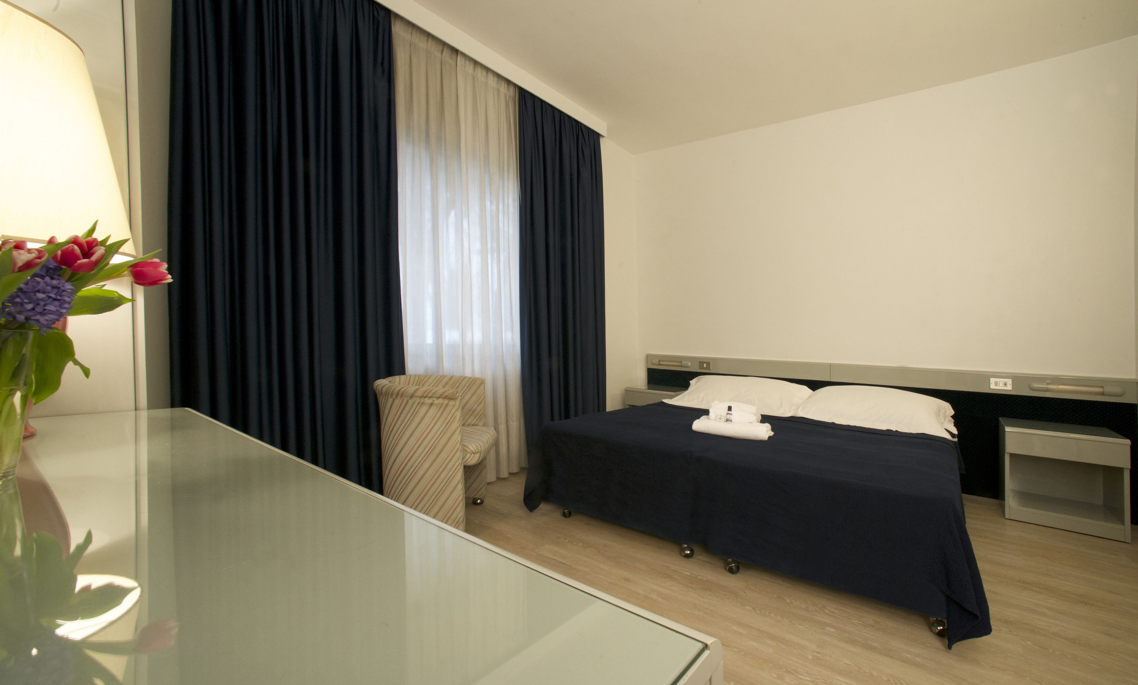 A hotel room with a bed, bedside table, lamp, and a vase with flowers.
