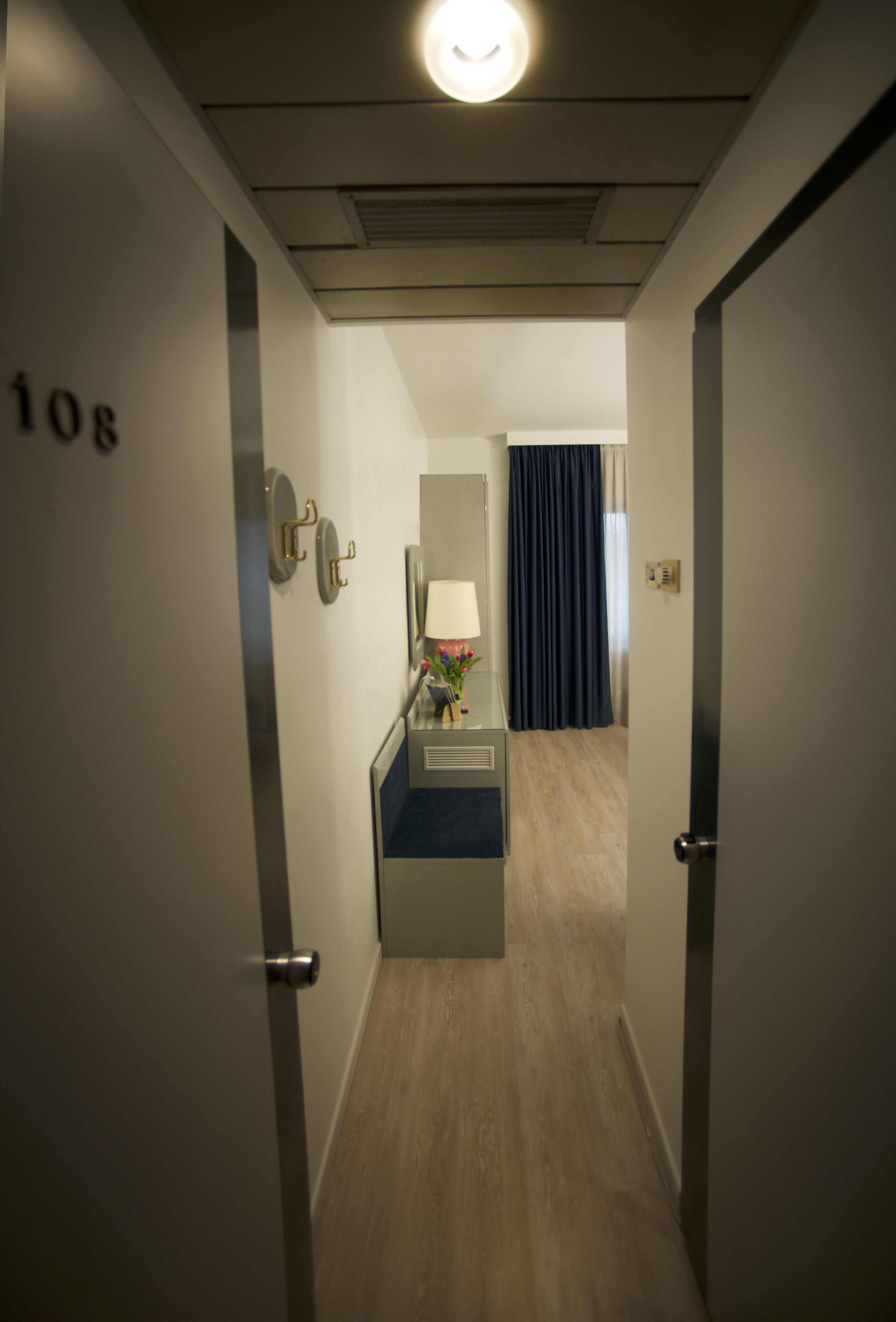 A hotel hallway with a number "108" on the wall, a small desk with a lamp, and a blue curtain in the background.