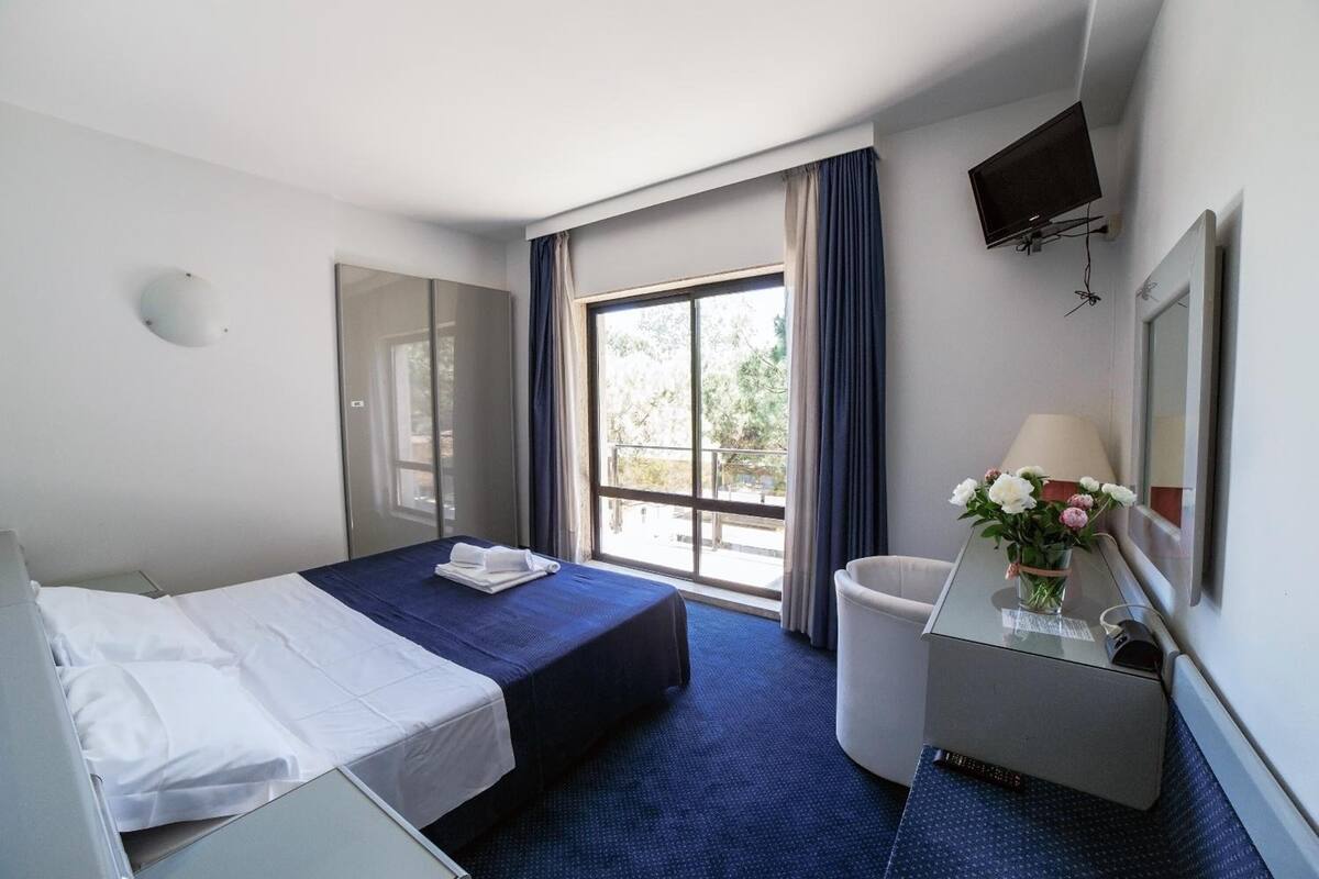 Une chambre d’hôtel avec un lit, un bureau doté d’une lampe, un téléviseur fixé au mur et une fenêtre avec des rideaux.
