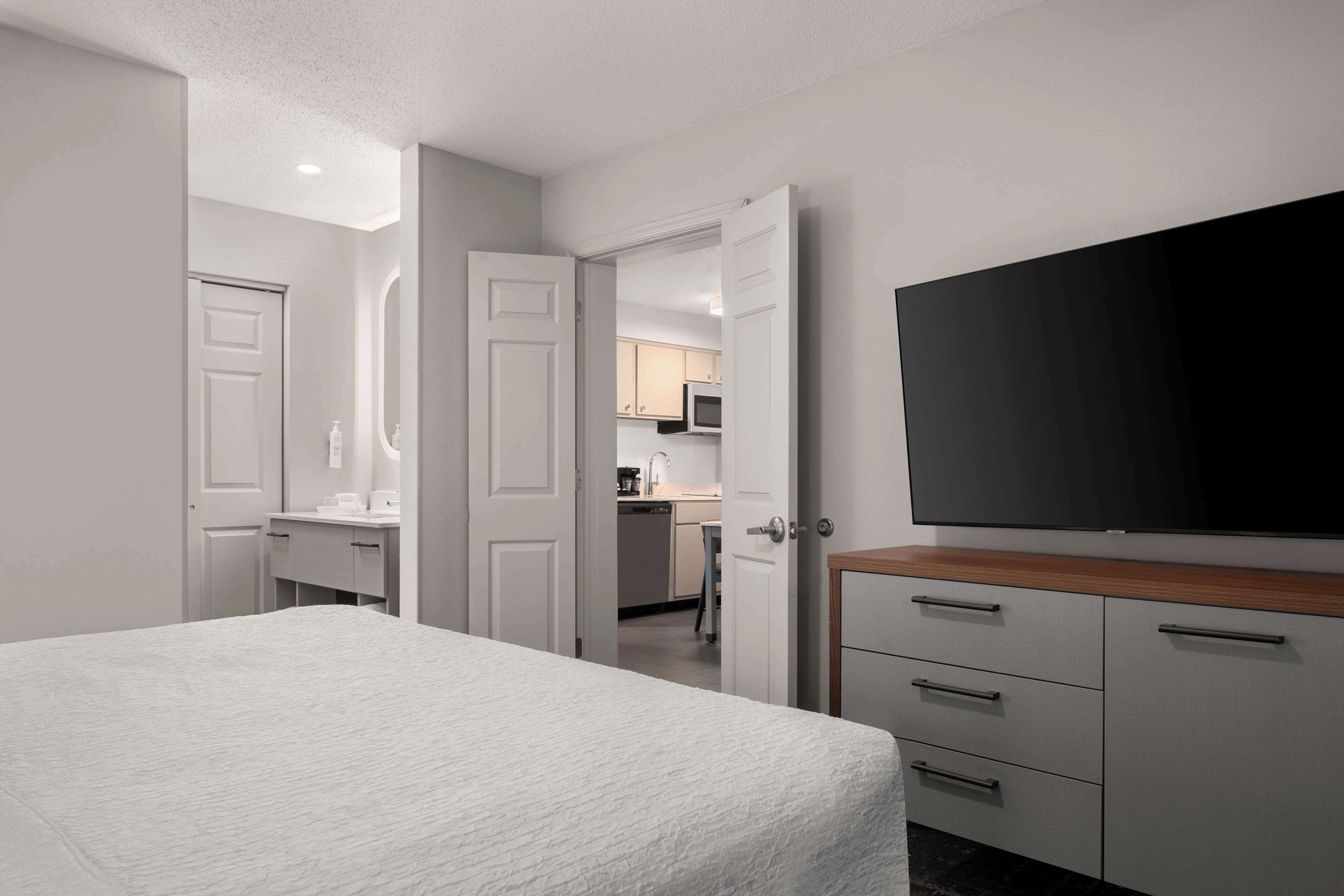 A bedroom with a large bed, a TV mounted on the wall, and a kitchenette in the background.