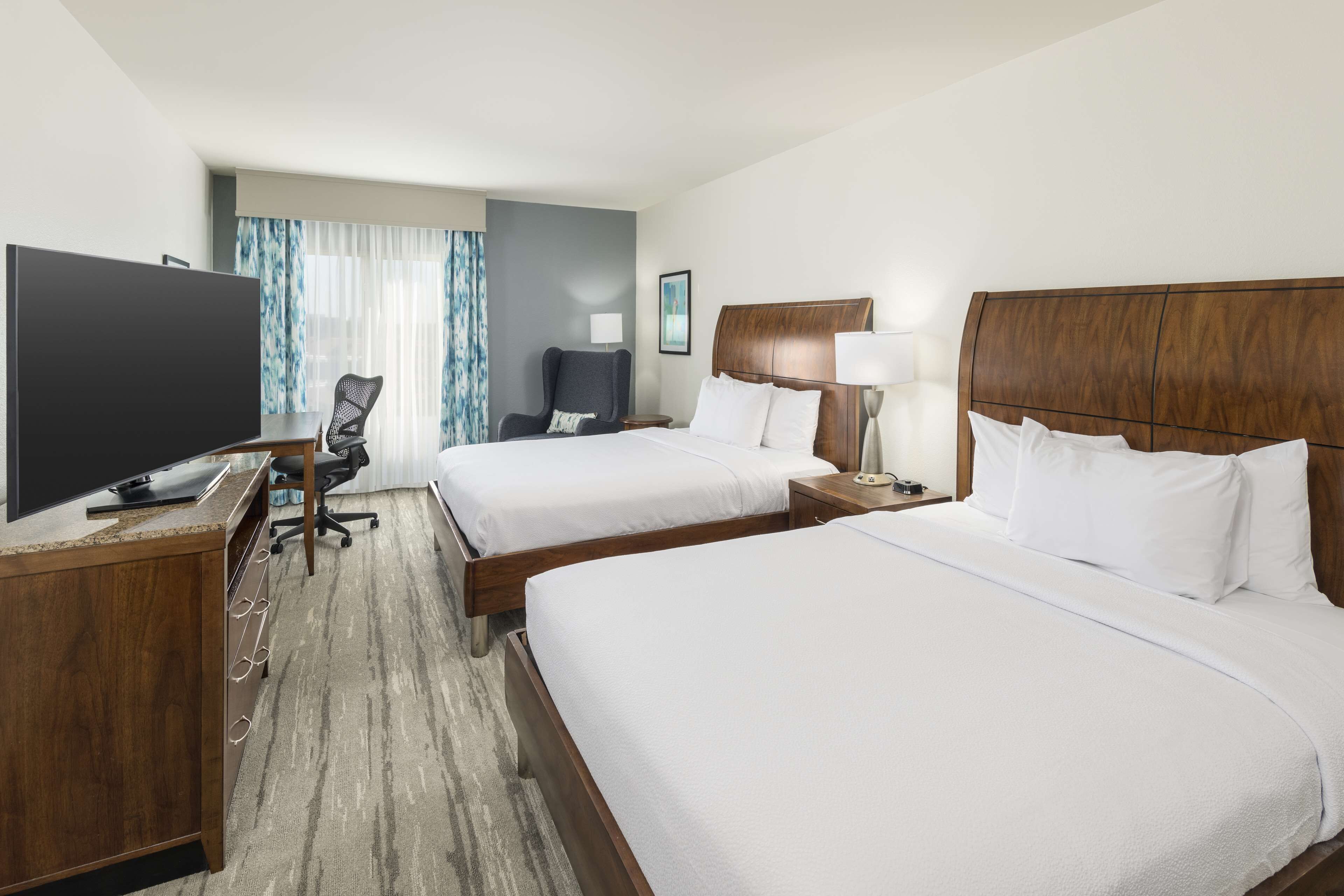 Quarto de hotel com duas camas, uma escrivaninha com televisão, uma cadeira e uma janela com cortinas.
