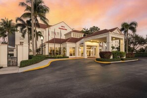 Hotel met overdekte ingang, palmbomen en een zonsondergang.