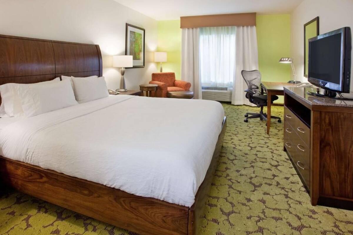 A hotel room with a large bed, a desk with a computer, a chair, a television, and a window with curtains.