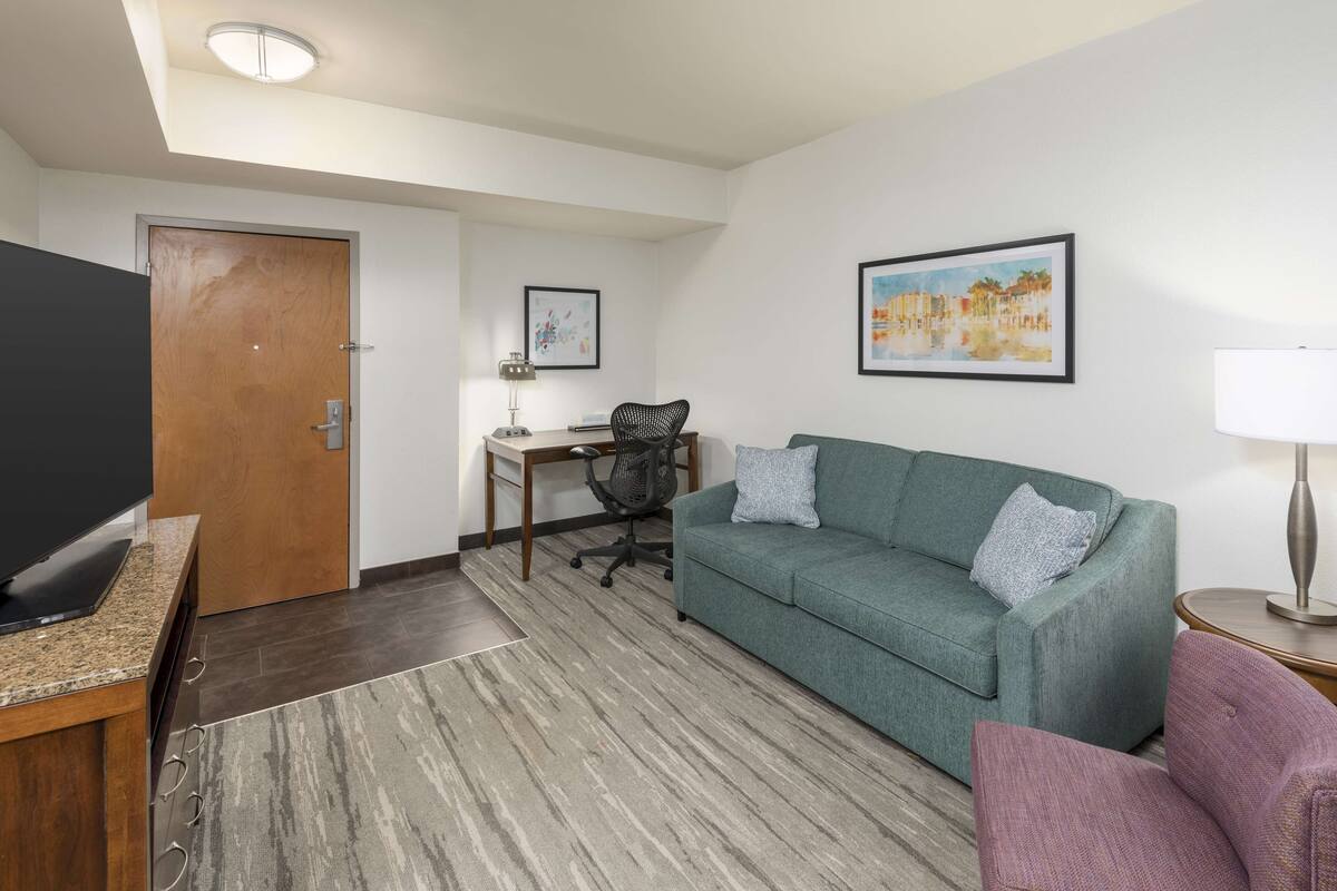 A modern living room with a green sofa, a flat-screen TV, a desk with a chair, and a painting on the wall.