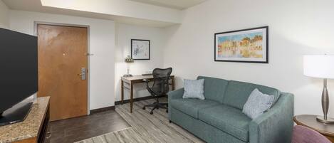 A modern living room with a green sofa, a flat-screen TV, a desk with a chair, and a painting on the wall.